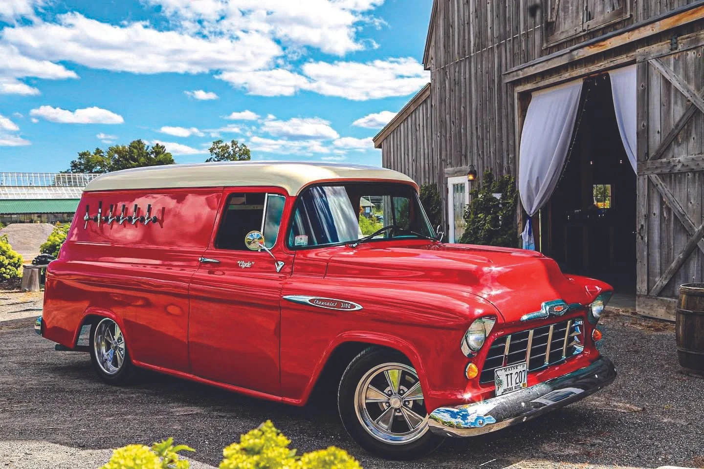 Red vintage Chevrolet mobile bar parked outside a rustic barn with white curtains, green plants and a blue sky with clouds.