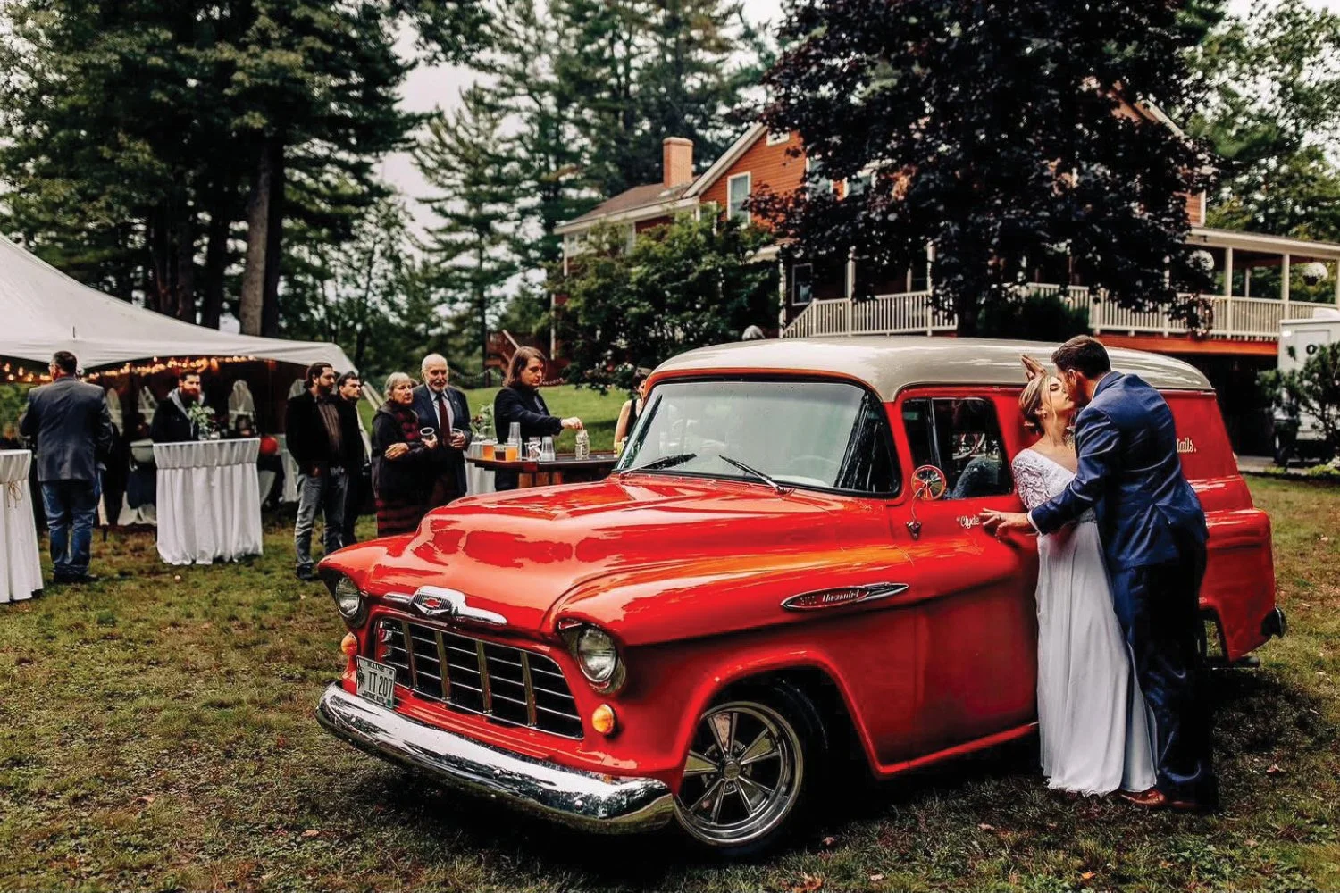 Couple kissing next to a vintage red Chevrolet truck at an outdoor wedding reception with guests and a white tent in the background.