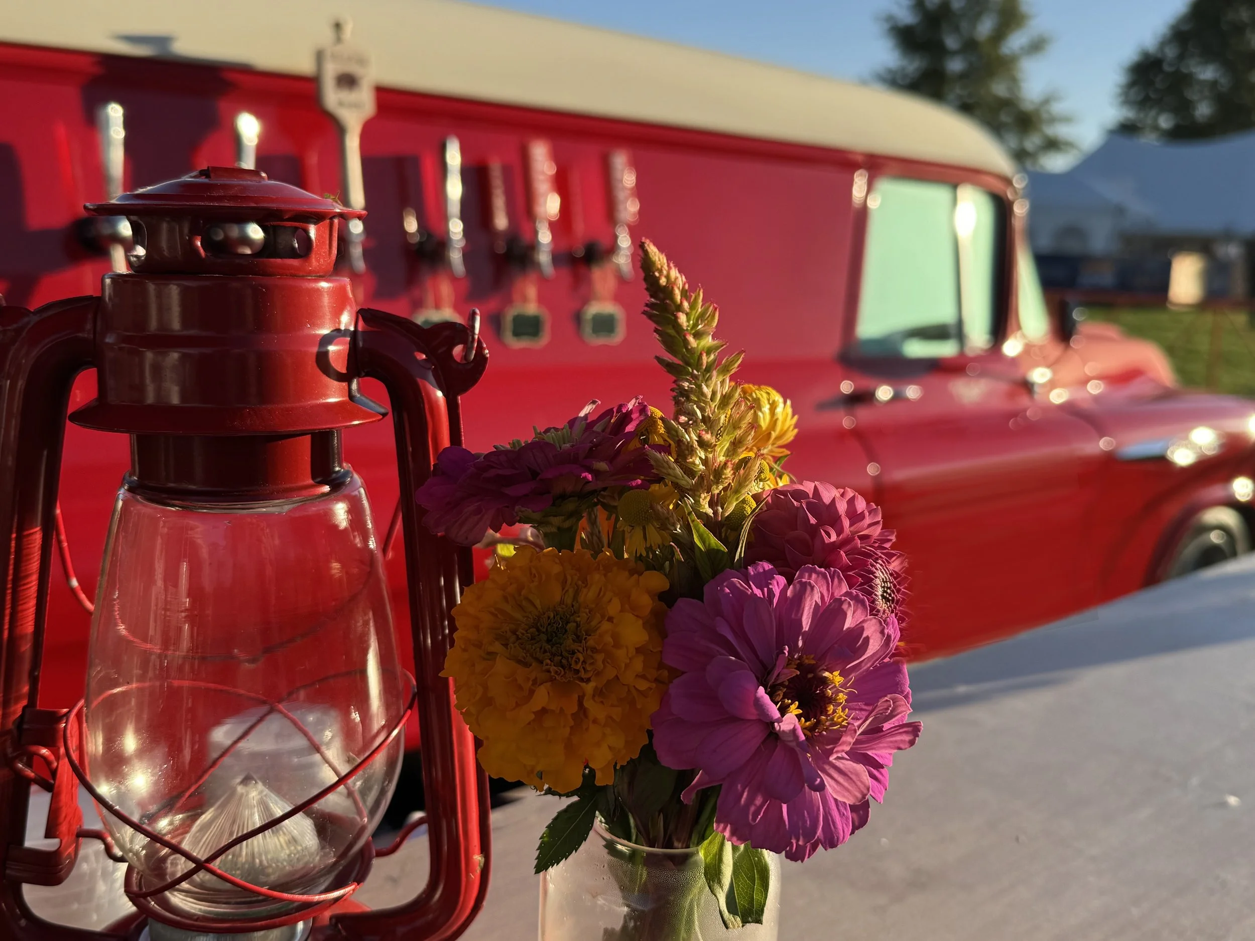 A red lantern and a bouquet of colorful flowers in a glass jar on a white surface, with a vintage red truck in the background under clear blue sky.