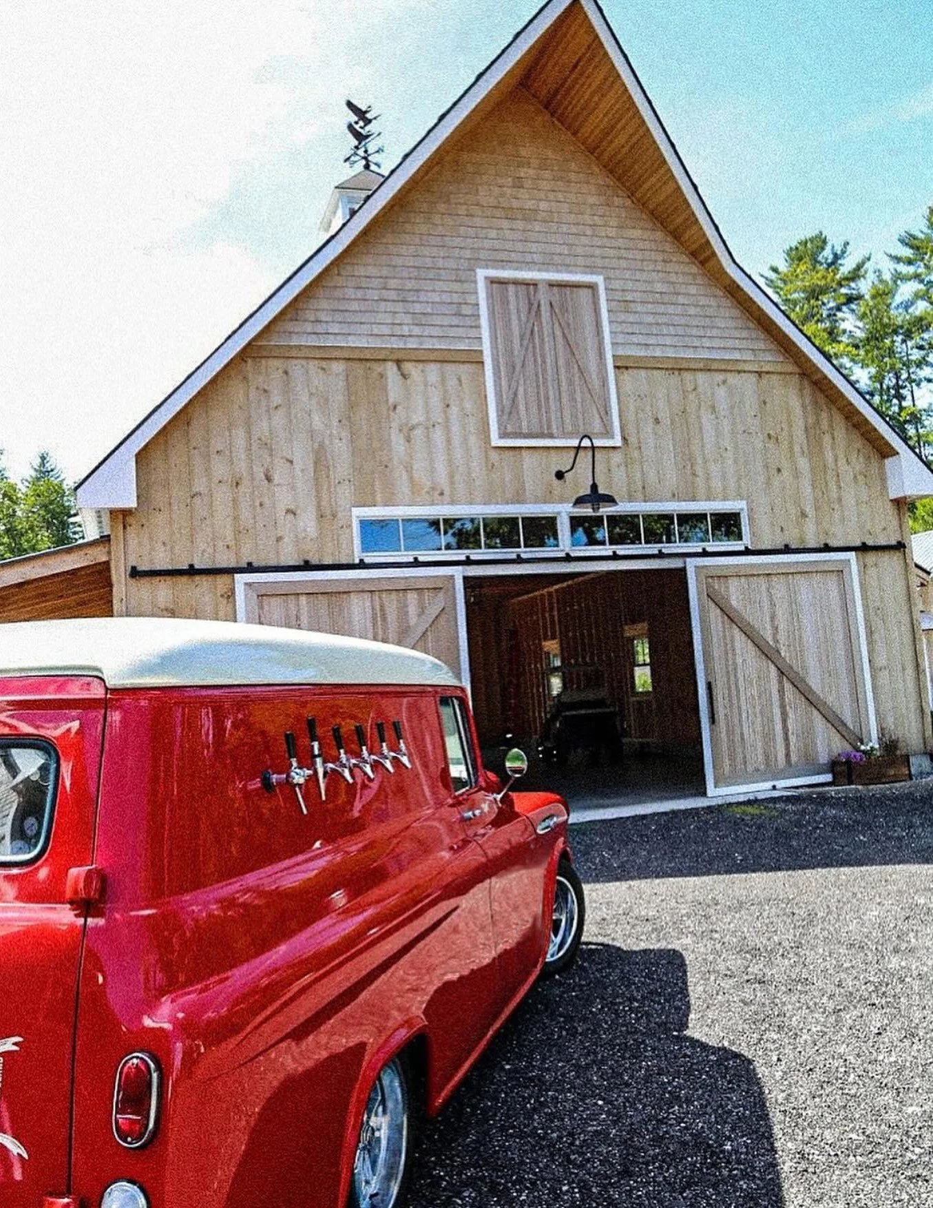 A red vintage Chevrolet mobile bar with a white roof parked in front of a newly built wooden barn with large sliding doors and a small upper window with shutters.