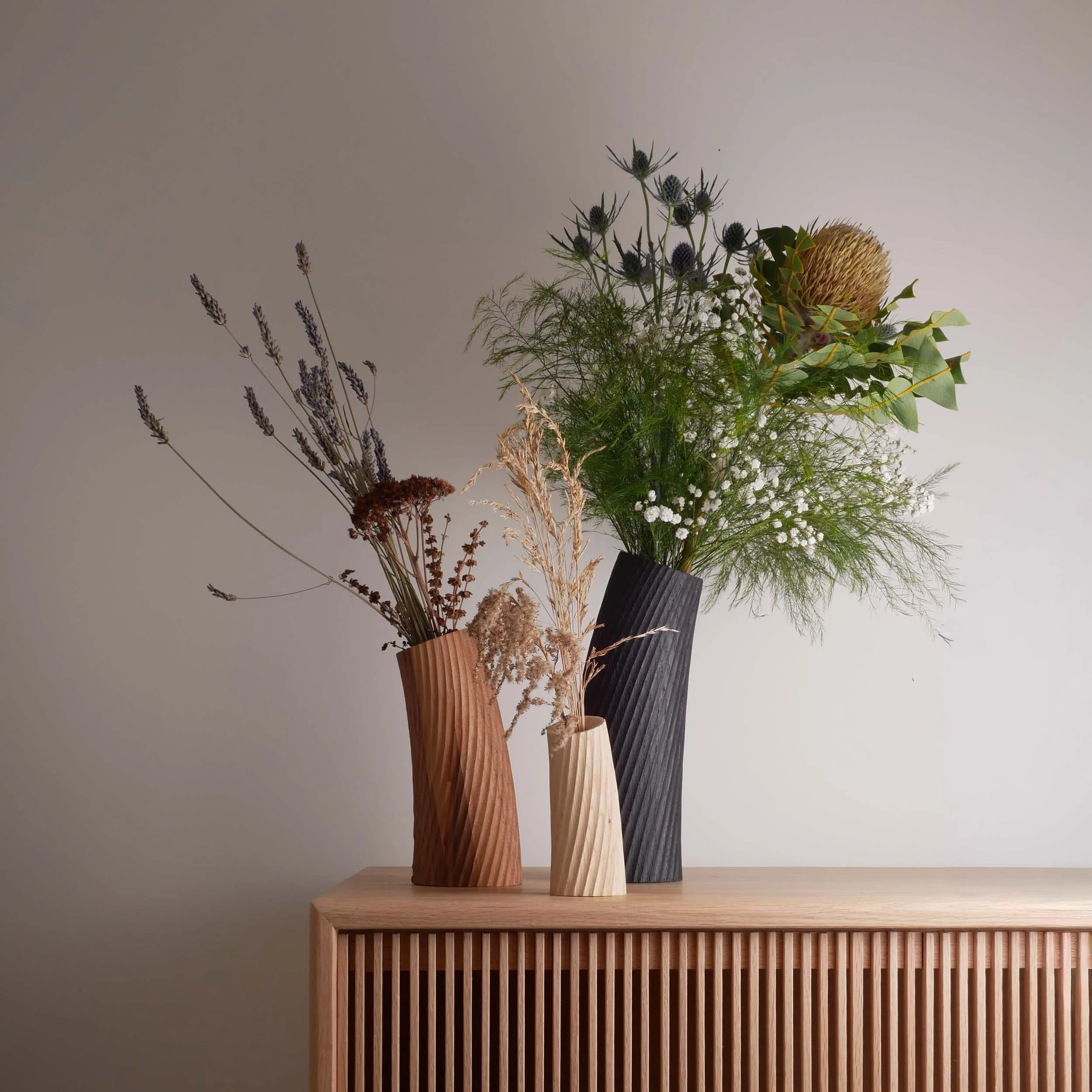 Three vases with dried and fresh flowers on a wooden sideboard against a plain wall.
