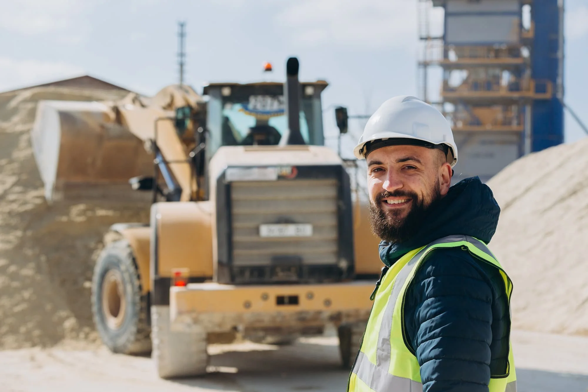 construction worker smiling with construction machine scooping in the background.