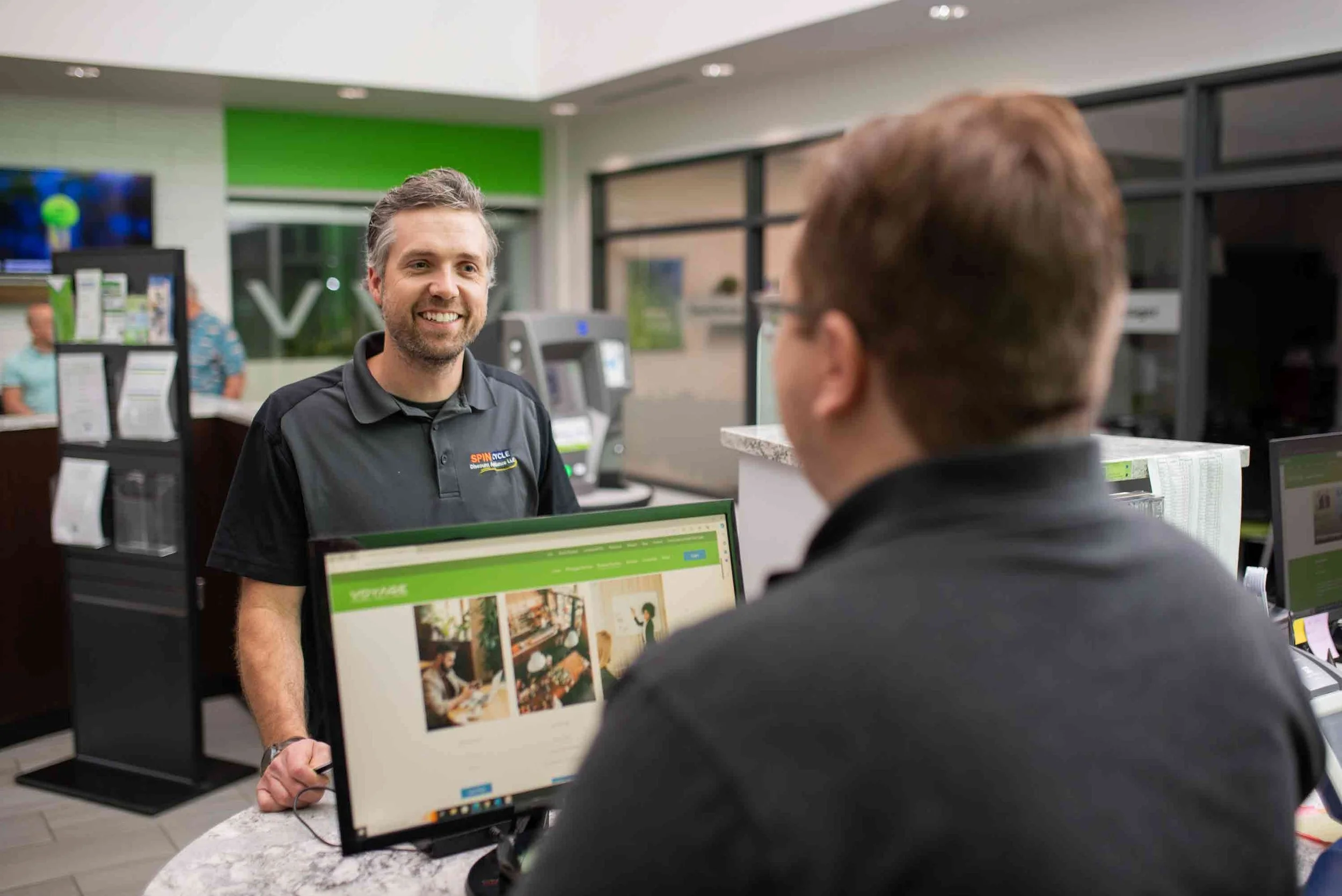 A man smiling at a customer service counter in a store, with a computer monitor displaying images of interior design and furniture, and a green and white color theme in the background.