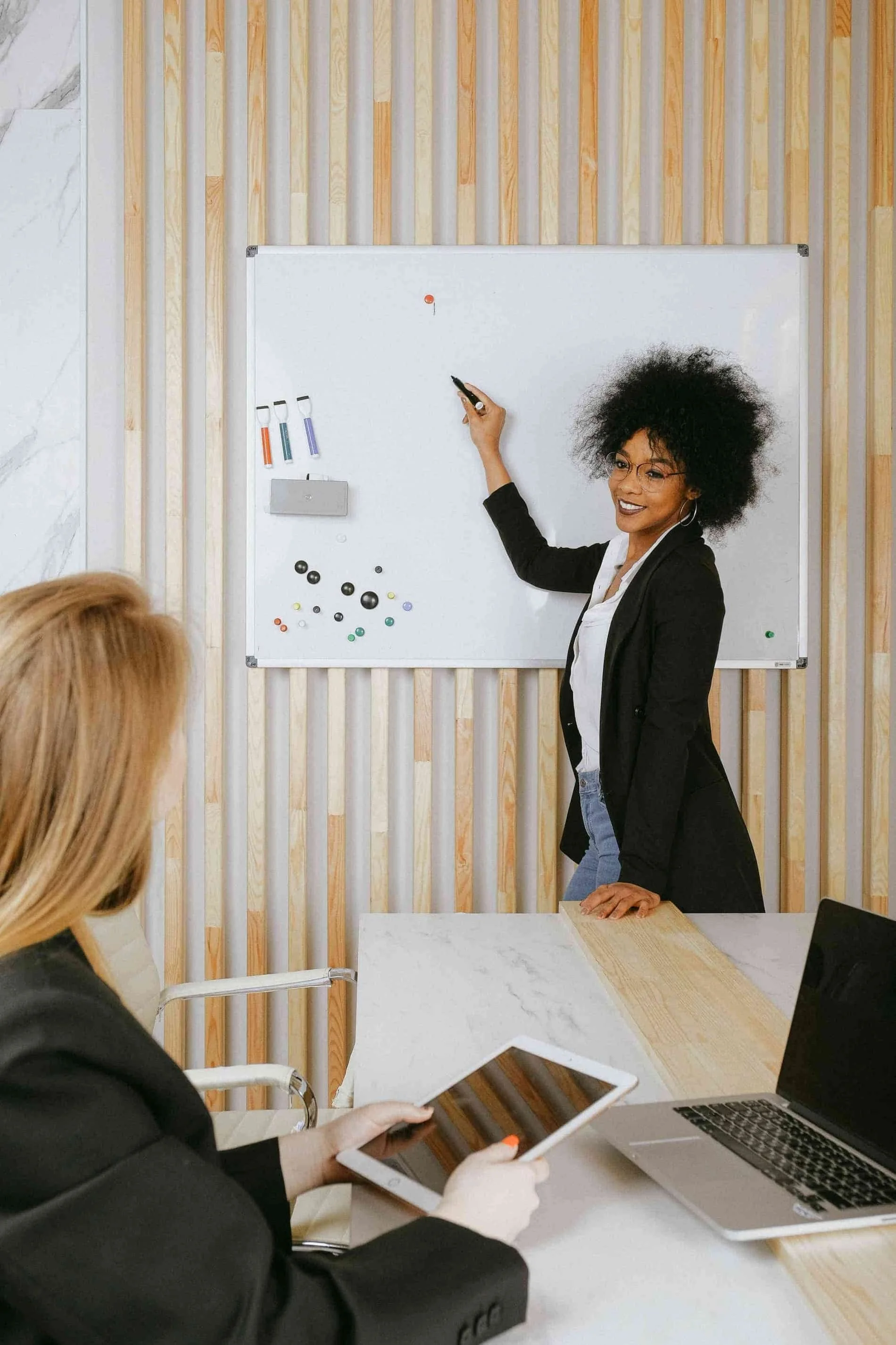 two ladies in a meeting, one holding an iPad, one writing on a white board