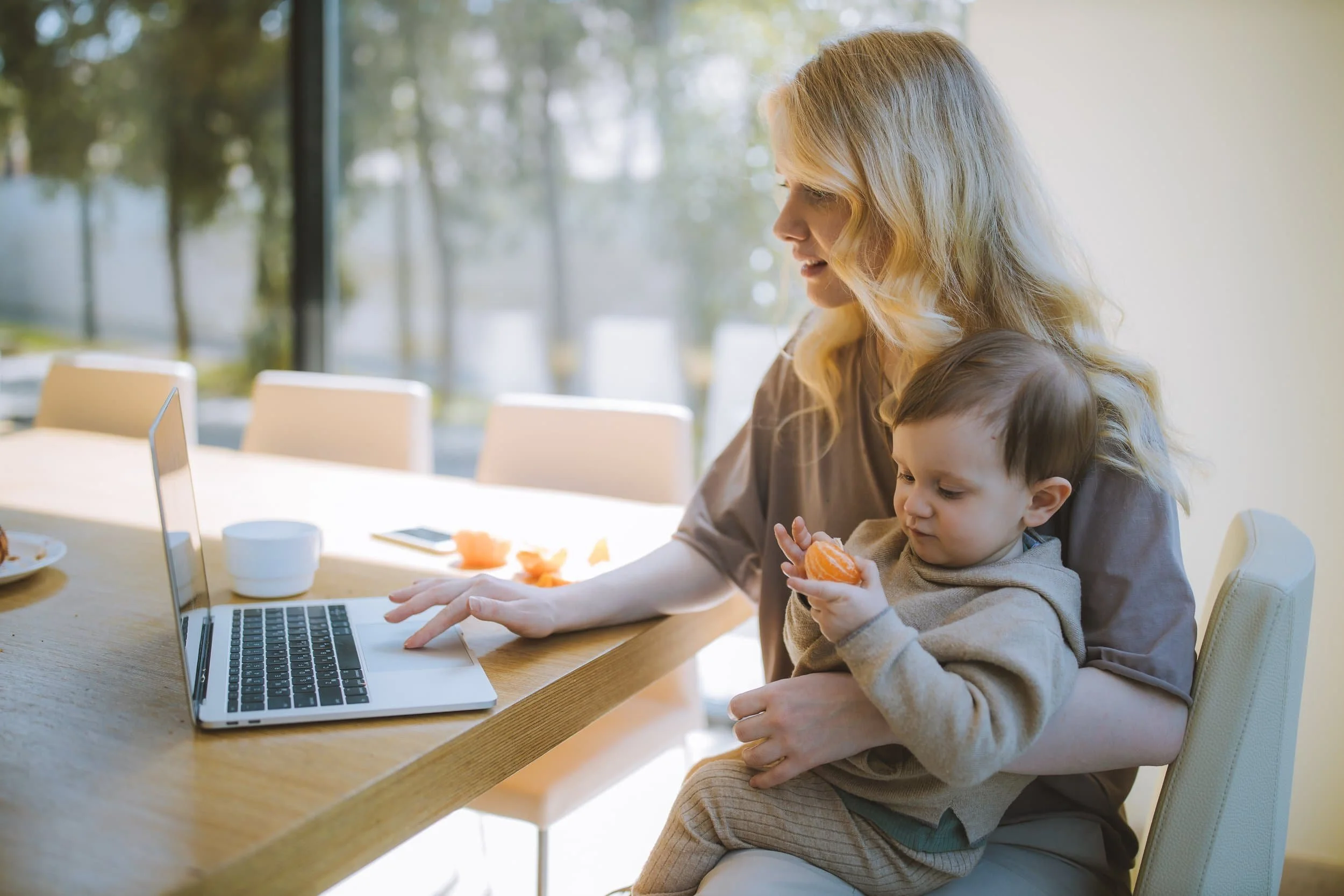 mother holding toddler at kitchen table working on a laptop
