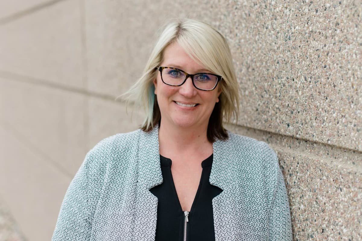 A smiling woman with blonde hair and glasses leaning against a textured beige wall outdoors.