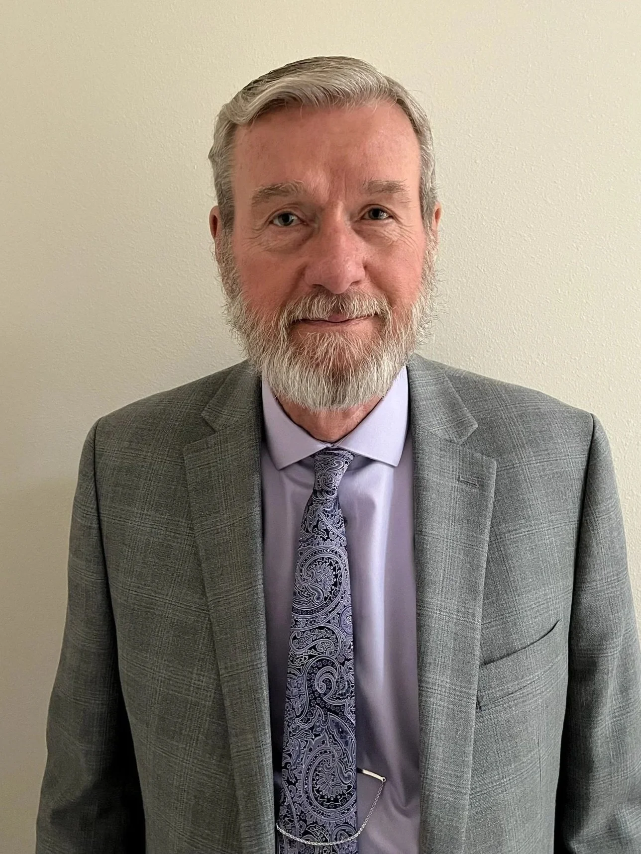 Portrait of a middle-aged man with gray hair, beard, wearing a gray checkered suit, a light purple shirt, and a paisley tie, standing against an off-white wall.