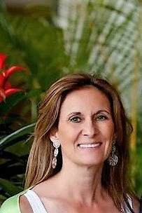 A woman with shoulder-length brown hair, wearing earrings and a sleeveless top, smiling outdoors with green plants in the background.