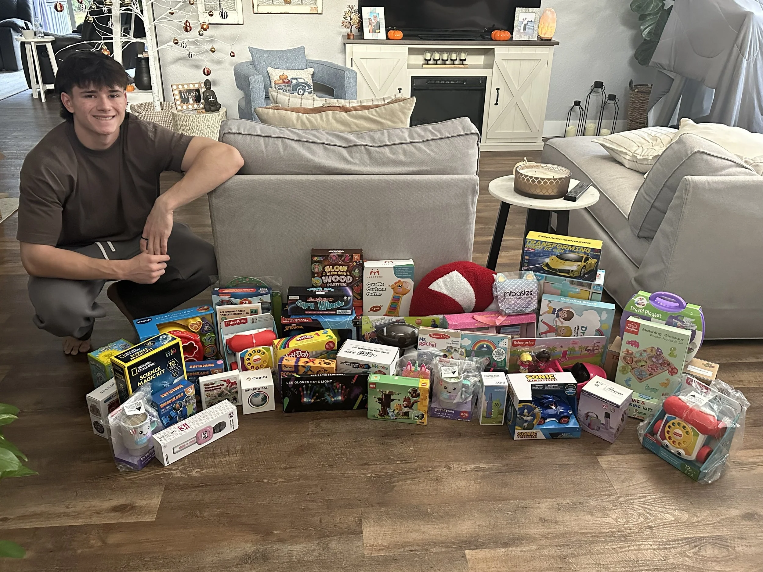 A young man with dark hair crouching on the floor next to a table filled with various toys and games. The background shows a cozy living room with a blue armchair, a white wooden sideboard decorated for Halloween, and a TV. The toys and games include educational kits, plush toys, and remote-controlled cars.