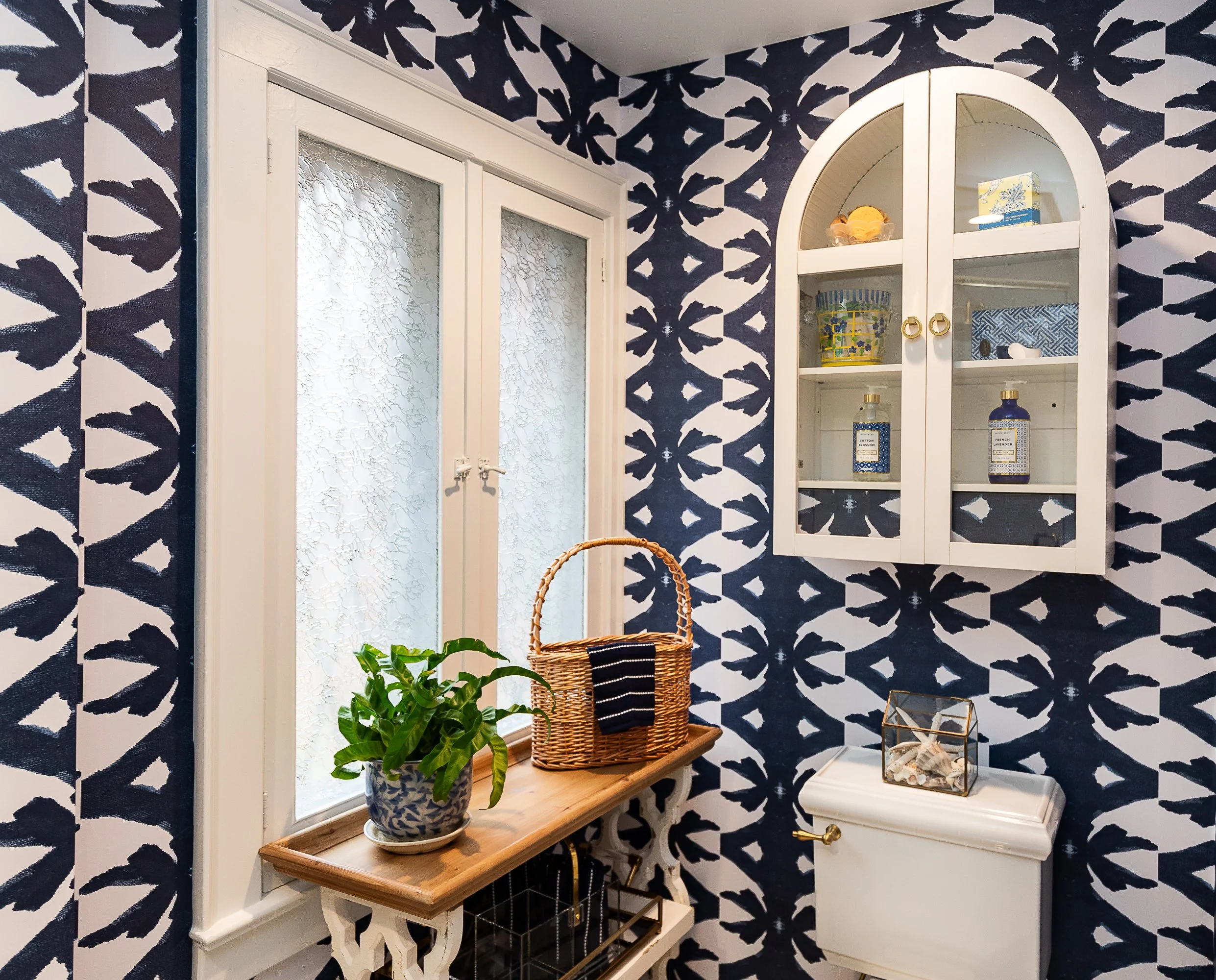 Decorative bathroom with navy and white patterned wallpaper, a white cabinet with glass doors, a wooden bench, a potted plant, and a glass terrarium on a white toilet tank.