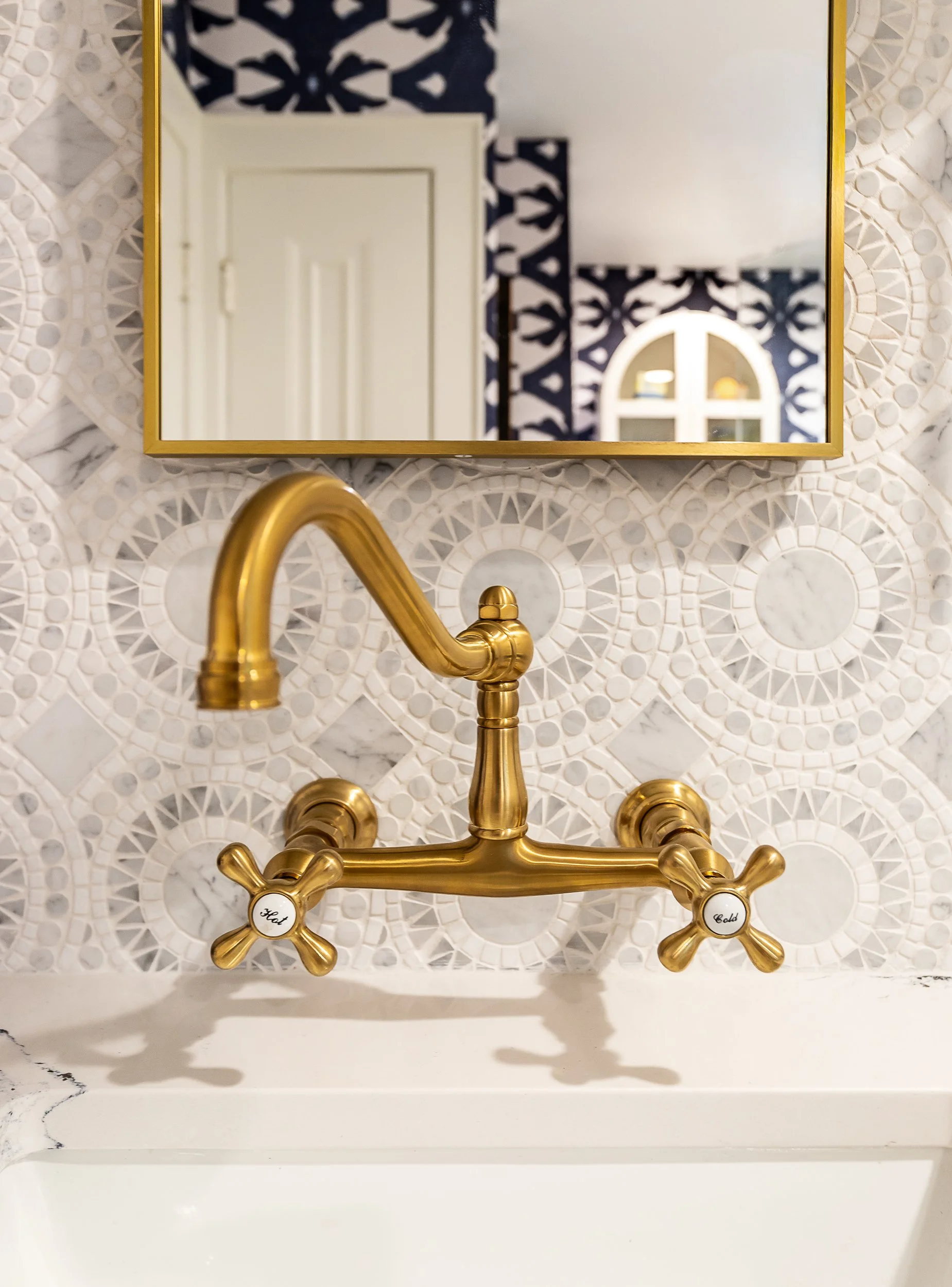 A gold vintage-style faucet mounted on a white marble countertop in a bathroom with a decorative geometric tile backsplash and a mirror reflecting a patterned wall and a window.