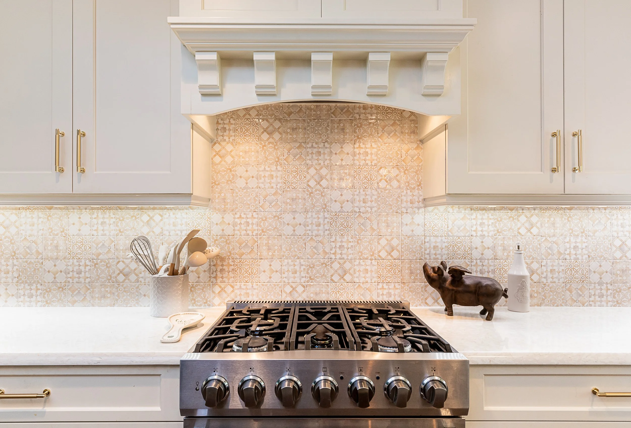 Kitchen countertop with a stove, utensil holder, and decorative pig figurine, with a patterned tile backsplash and beige cabinetry.