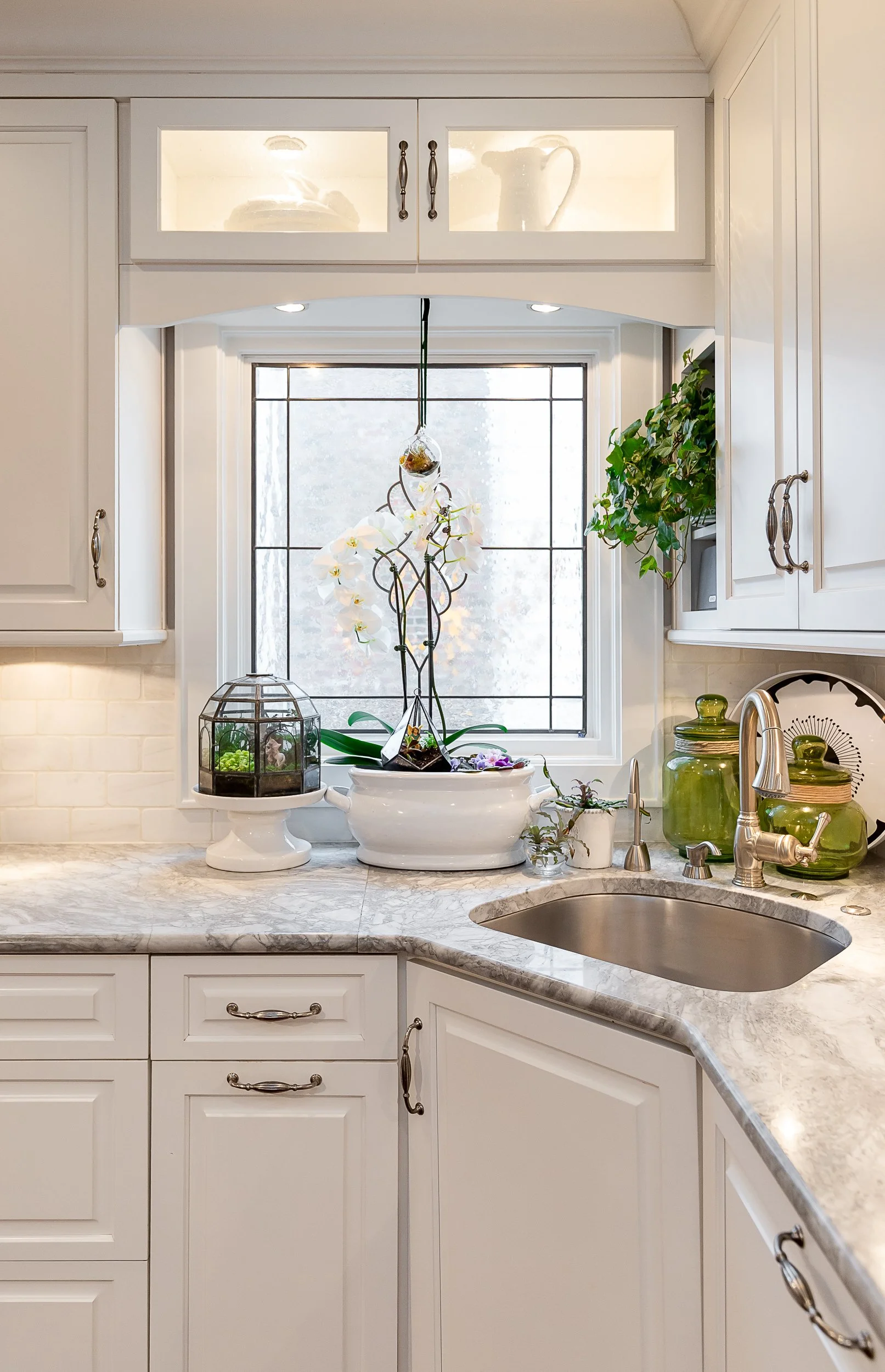 A kitchen with white cabinets, a marble-looking countertop, a sink, and a window decorated with orchids and various plants and decor pieces.