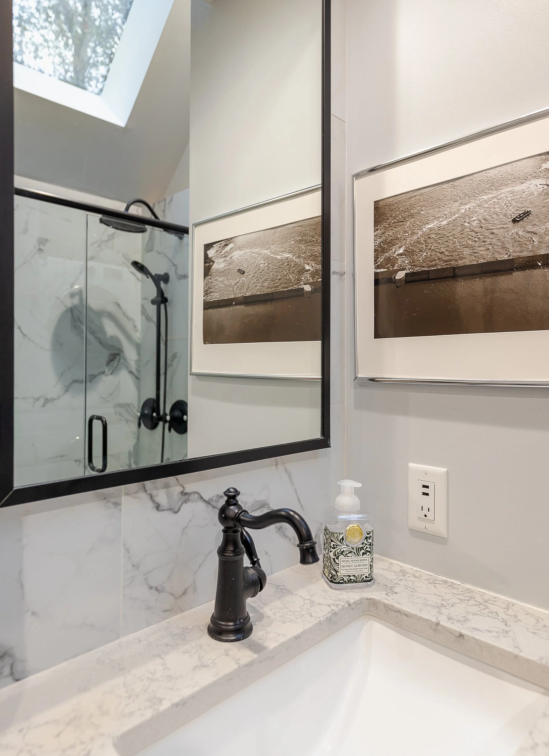 Bathroom sink with marble countertop, black faucet, mirror, framed artwork, soap dispenser, electrical outlet, shower area with glass door, and skylight.