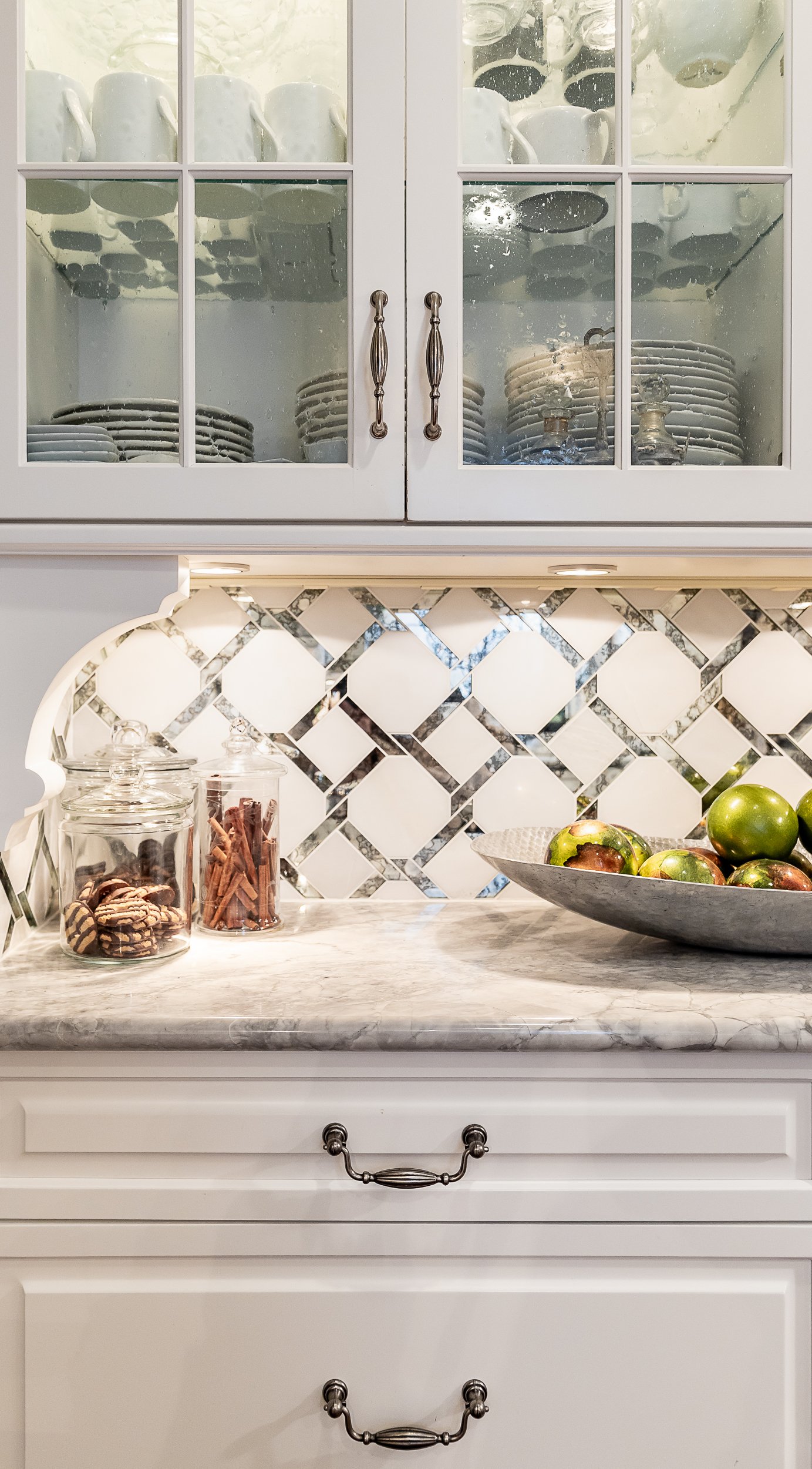 Kitchen cabinet with glass doors containing plates, cups, and glassware, quartz countertop with jars of cookies and cinnamon sticks, bowl of green apples, and decorative backsplash tiles.