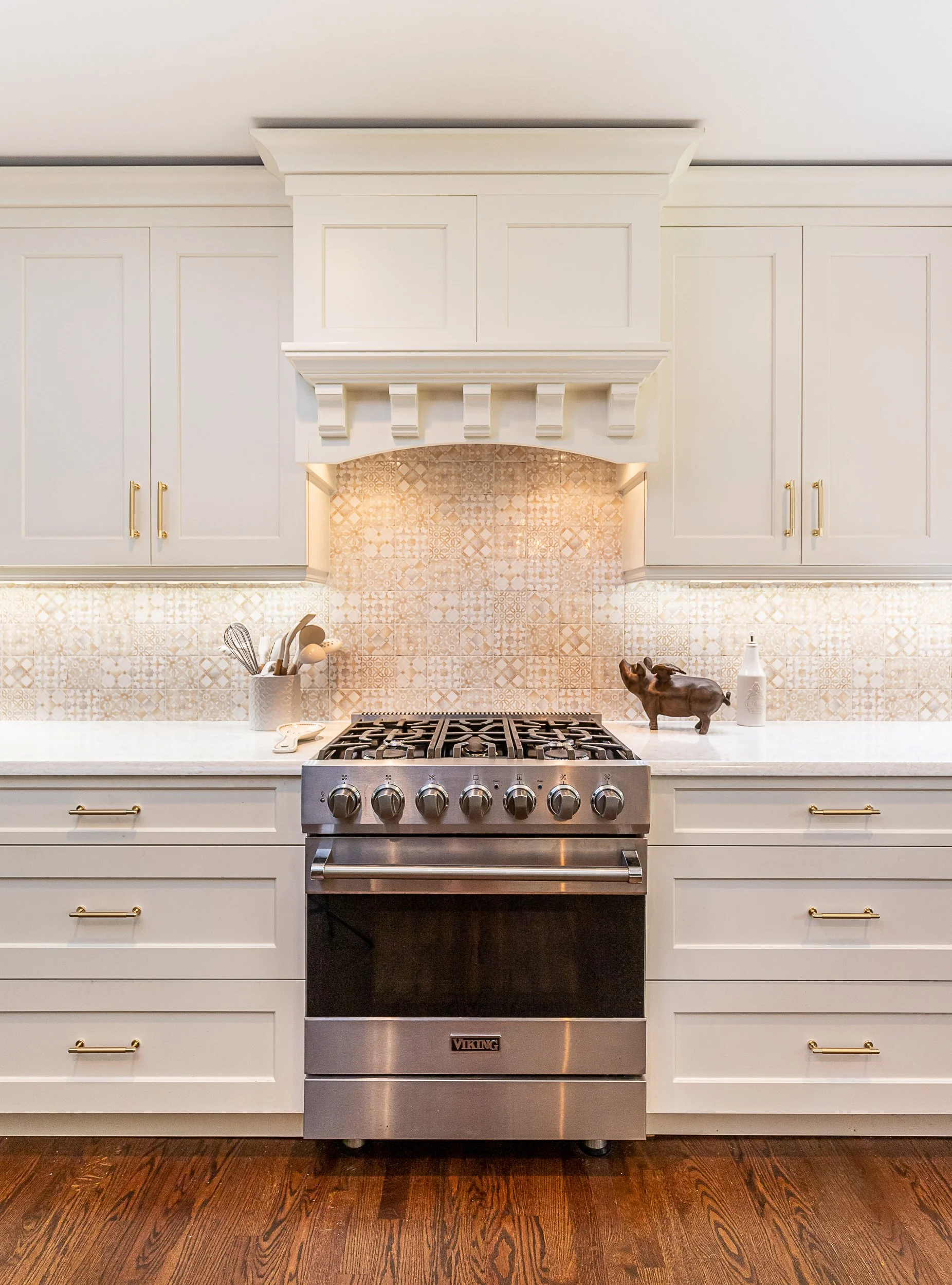 Kitchen with a stainless steel Viking oven, Beige cabinets with gold handles, beige patterned backsplash, and decorative items on the countertop including a ceramic pig.