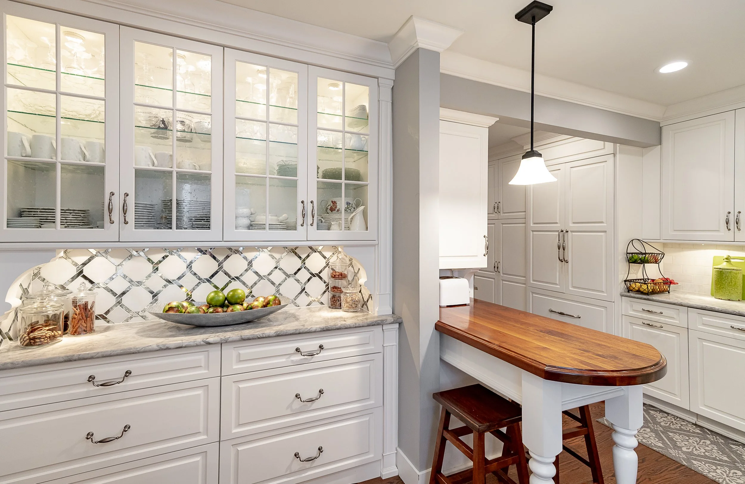 Modern kitchen with white cabinetry, a quartz countertop, glass-front upper cabinets, a small wooden breakfast table with a stool, and a pendant light. Items include a fruit bowl with green apples, jars with snacks, and a wire basket with fruits.