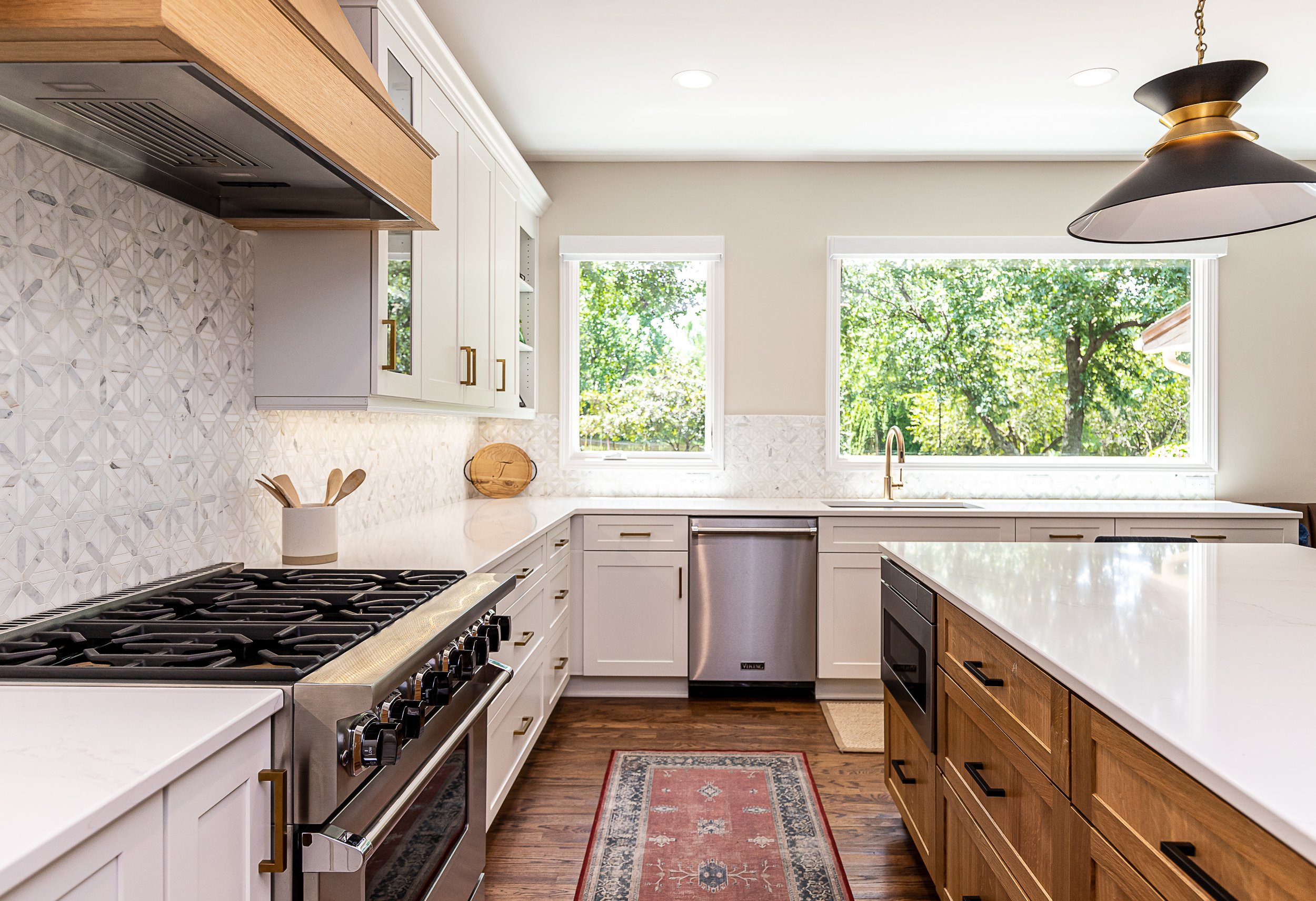 Bright kitchen with white cabinets, a wooden kitchen island, stainless steel stove and dishwasher, two large windows showing green trees, a rug on wooden floor, and modern black and gold pendant lighting.