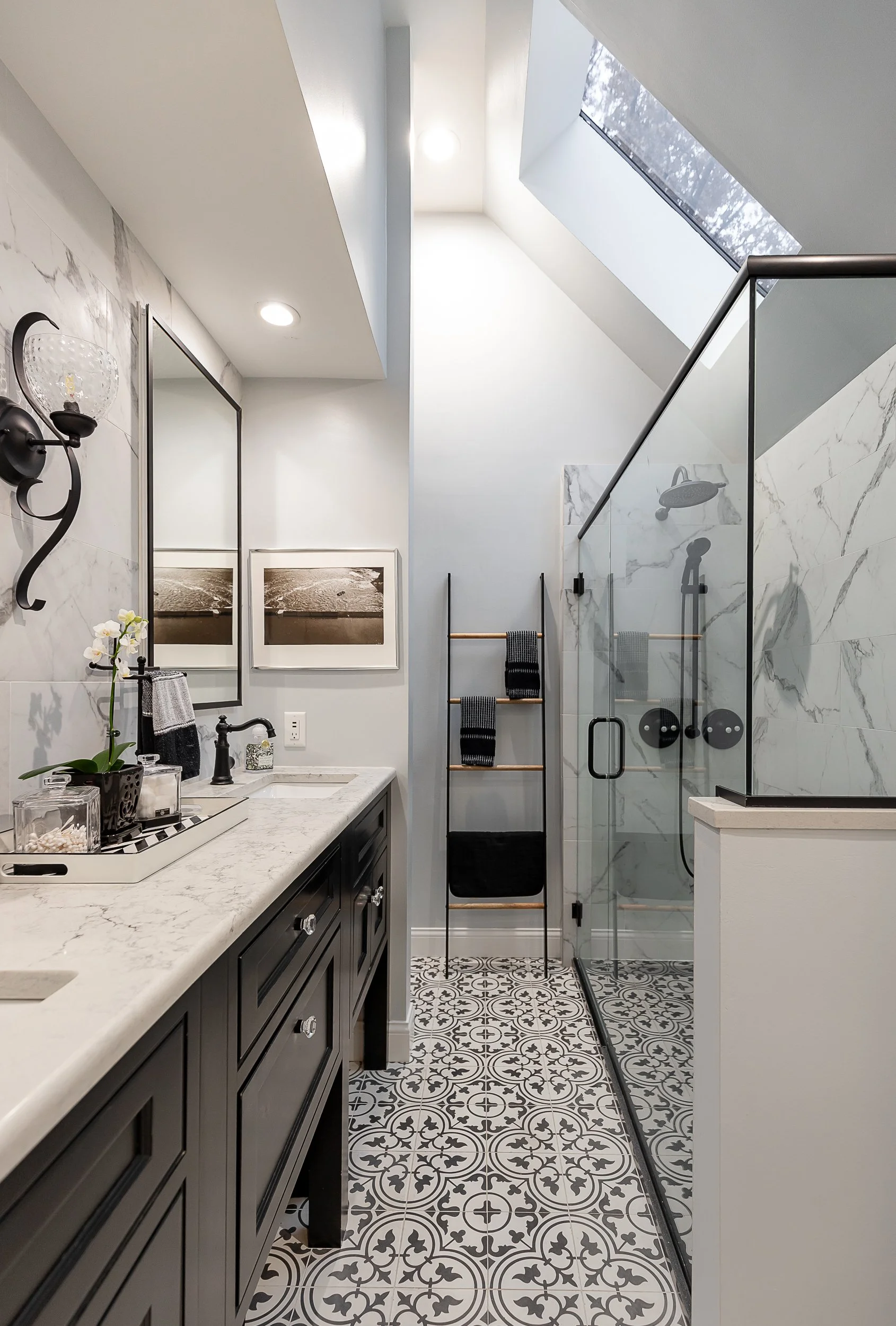 Modern bathroom with black vanity, white marble countertop, framed black and white photographs, wall-mounted sconce, towel rack with black towels, glass shower with marble tiles, patterned floor tiles, skylight, and black fixtures.