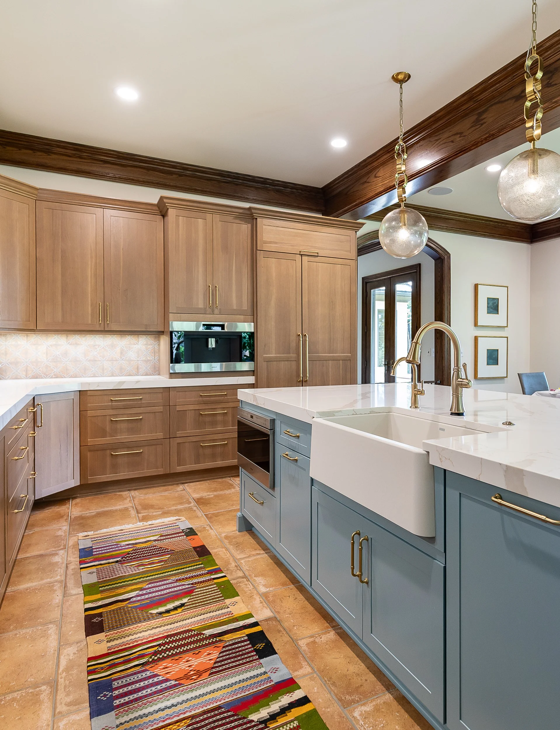 A modern kitchen featuring upper wooden cabinets with gold handles, a marble island featuring a farmhouse sink and gold fixtures, pendant lights with glass globes and gold chains, a doorway with a wooden frame leading to a backyard patio, wall art with framed pictures, a colorful striped runner rug on the tile floor.