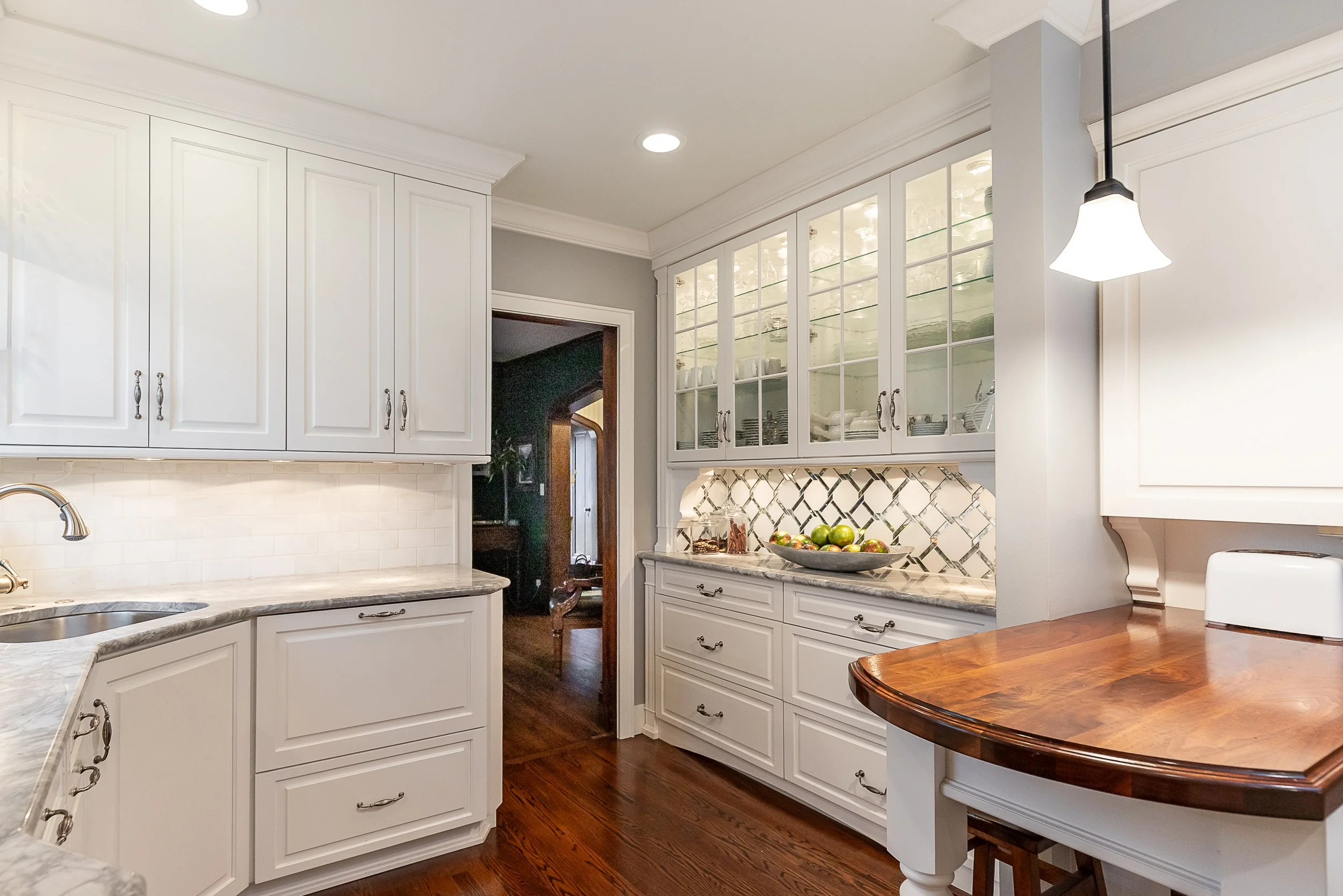 A kitchen with white cabinets, a marble-like countertop, a backsplash, a small eating nook, and a doorway leading to a darker room.
