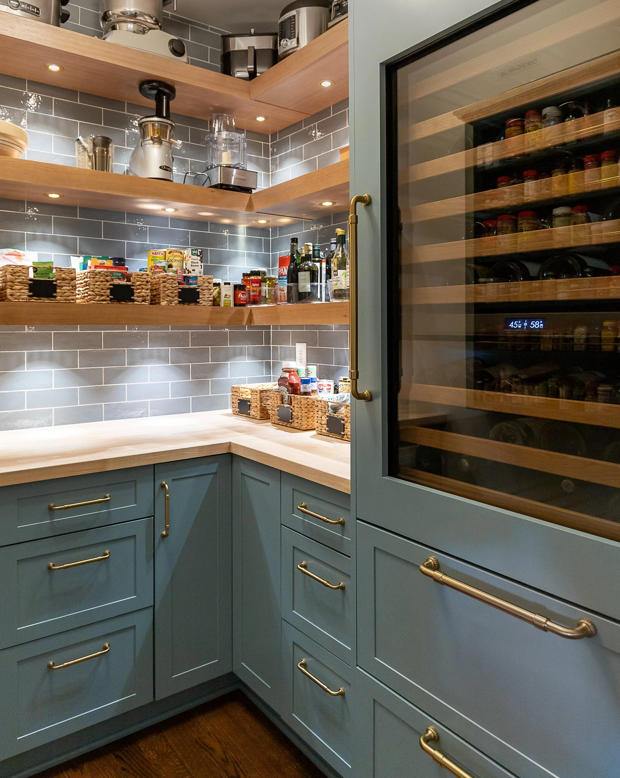 Kitchen with blue cabinets featuring gold handles, wooden open shelves, a glass-door wine fridge, gray tiled backsplash, and various food cans and bottles on the counter.