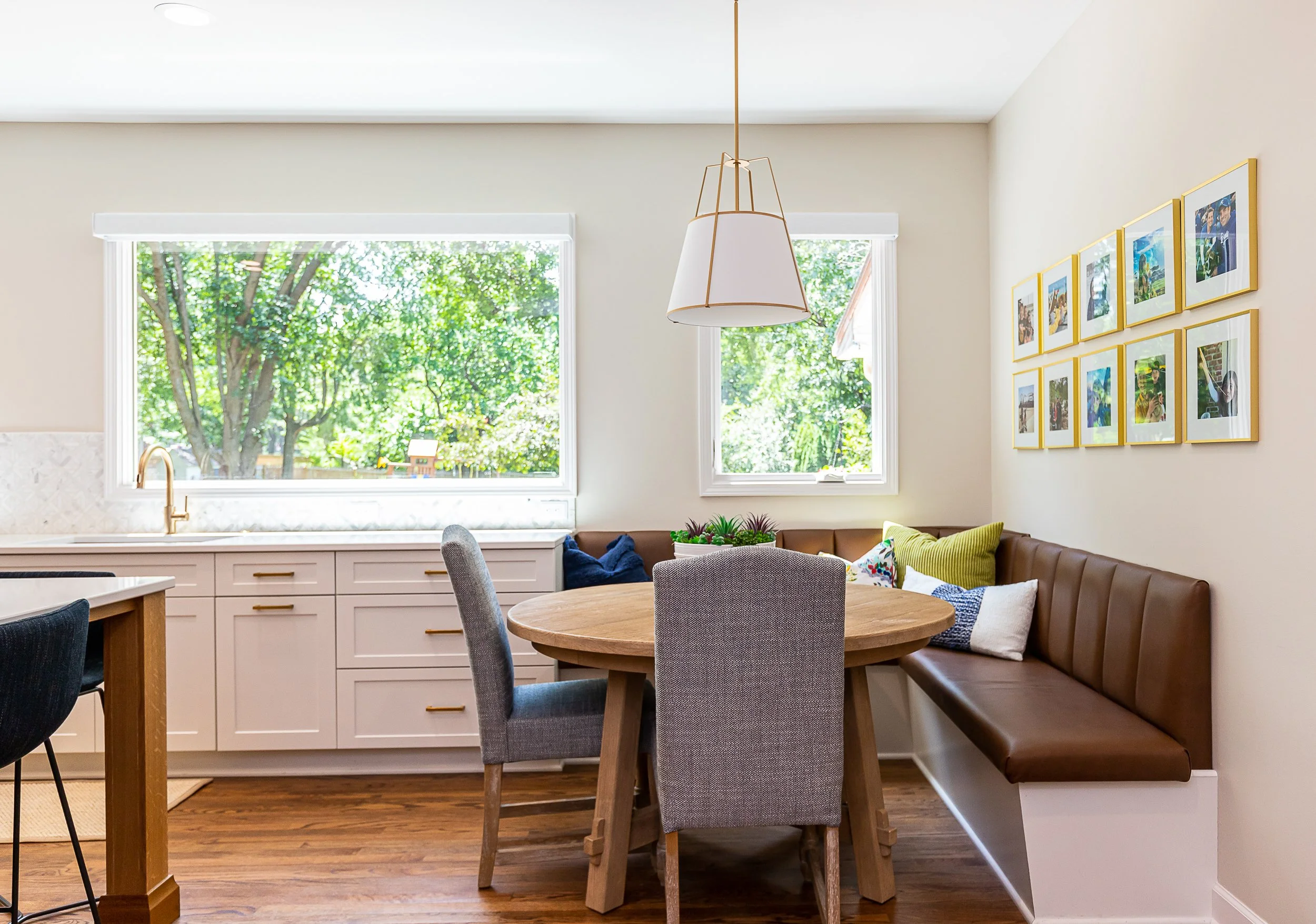 A cozy kitchen and dining area with large windows showing green trees outside, white cabinetry, a round wooden table, gray and black chairs, a brown cushioned bench with decorative pillows, and framed photos on the wall.