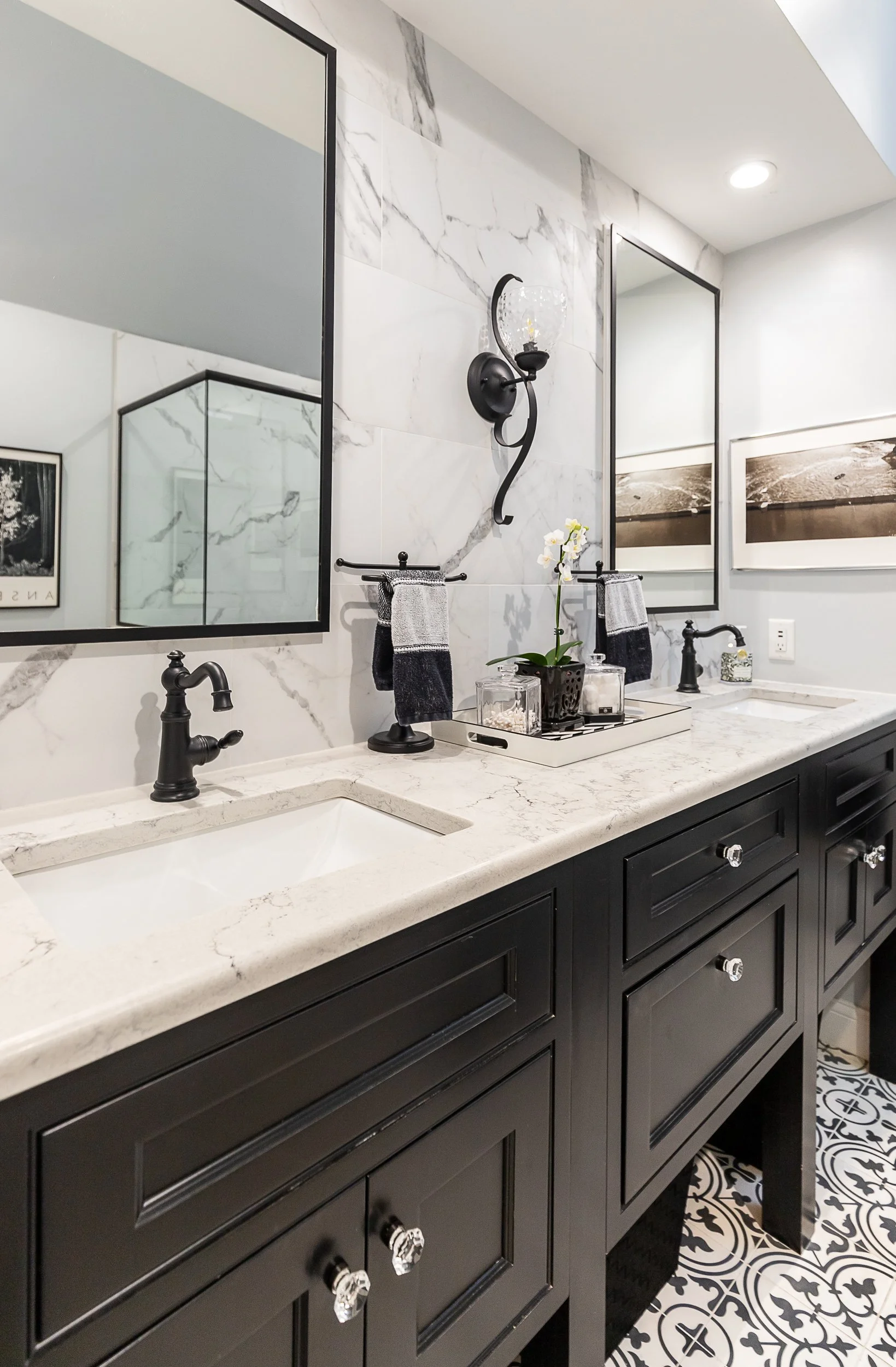 Bathroom vanity with dark cabinets, white marble-like countertop, two sinks, black fixtures, large mirrors, wall sconce light, decorative items including a potted orchid, and patterned black and white floor tiles.