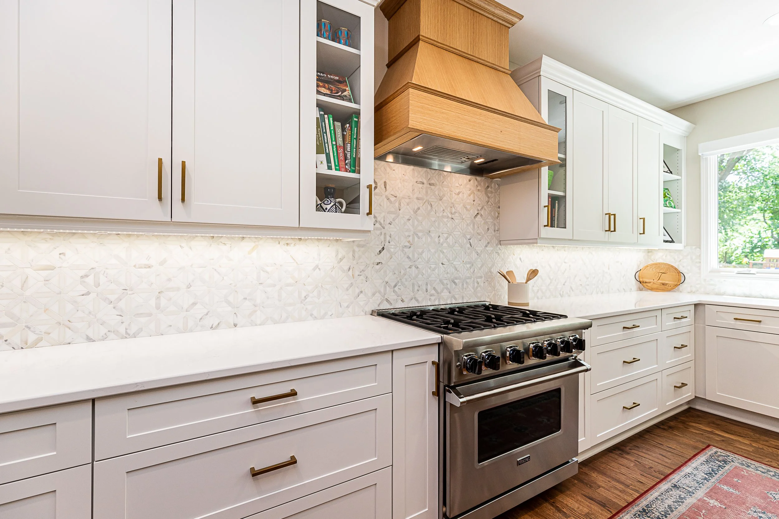White kitchen cabinets with gold handles, stainless steel stove, wooden range hood, patterned tile backsplash, and a window with green trees outside.