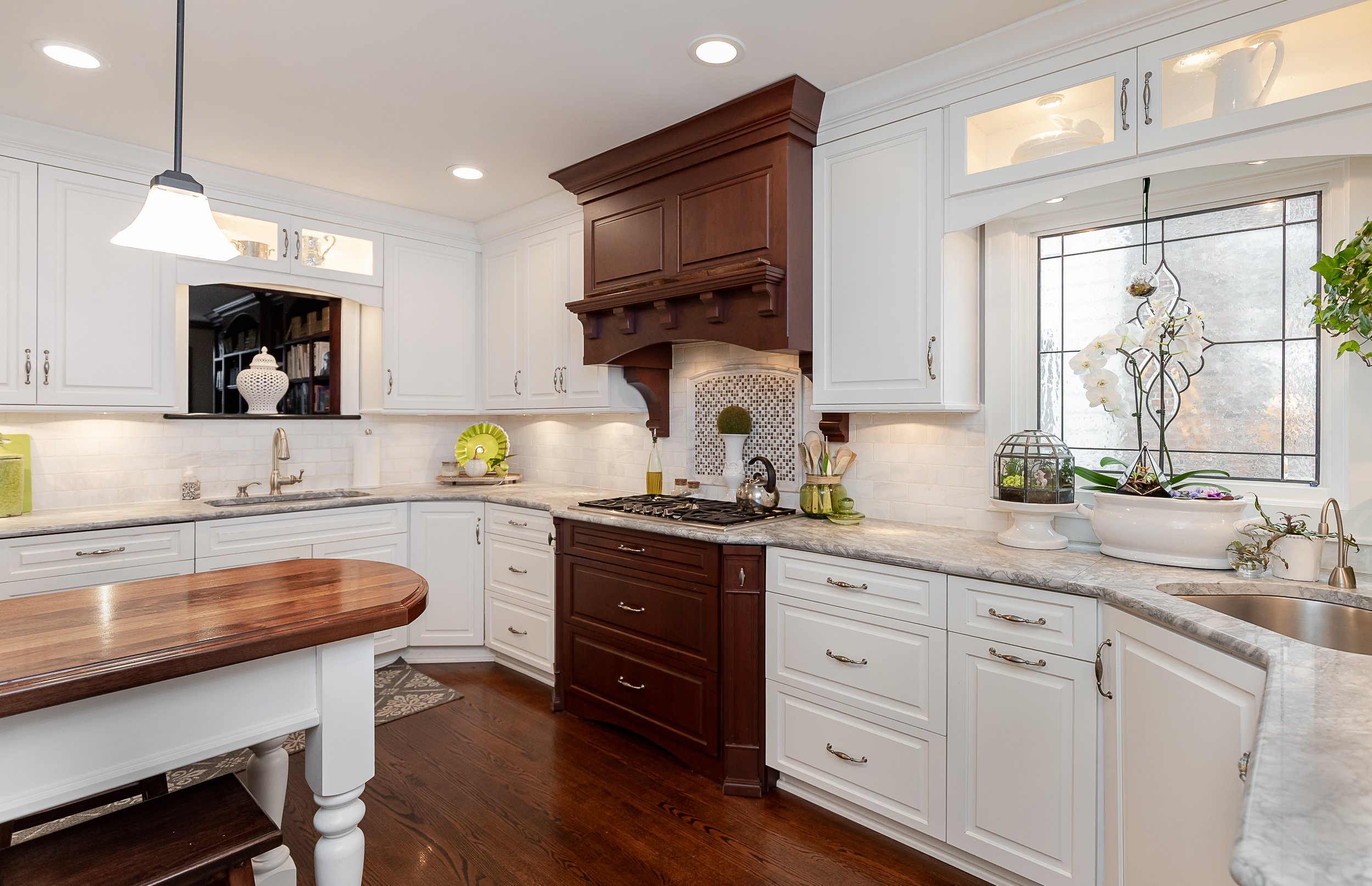 Kitchen with white cabinets, quartz countertops, a wooden island, a brown range hood with decorative molding, and a window with a plant and decorative items.