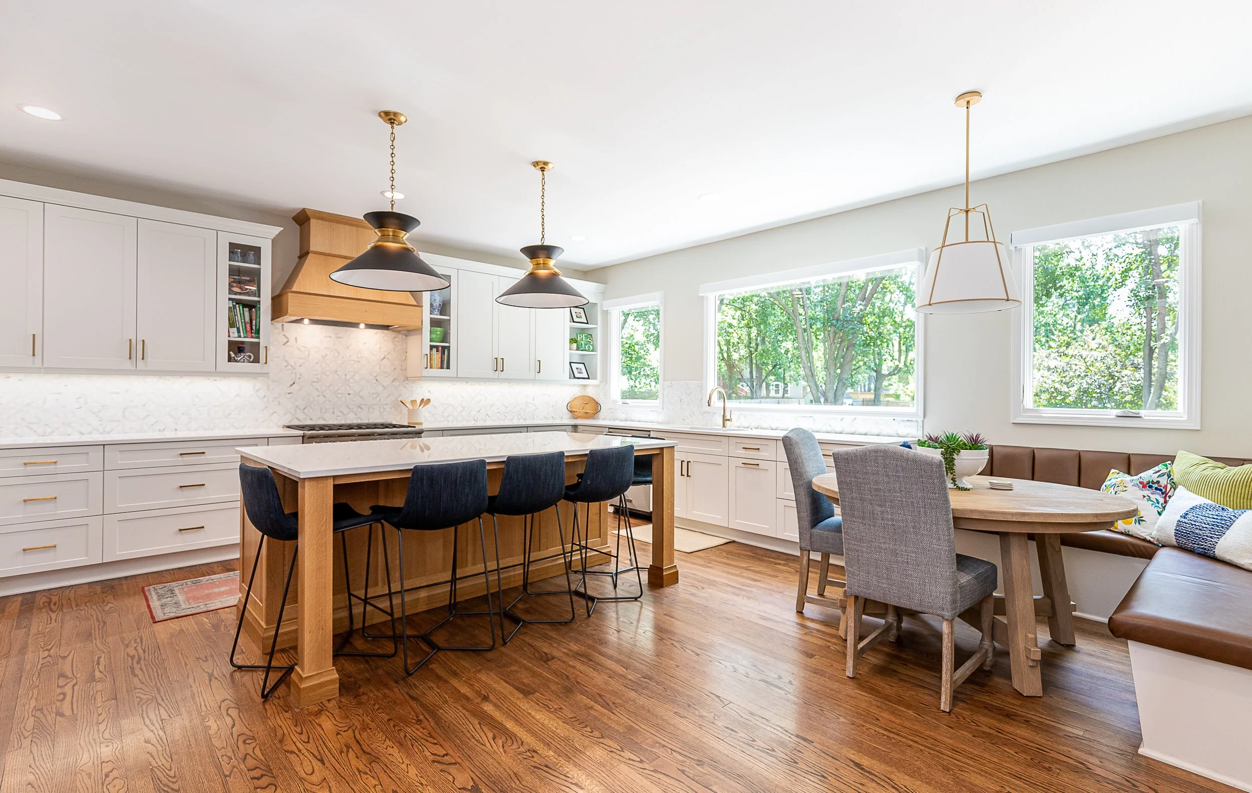 Bright kitchen with white cabinets, wooden accents, and hardwood flooring, featuring a kitchen island with four black chairs, a dining area with a round table and gray and green seating, large windows with a view of green trees outside, and modern pe