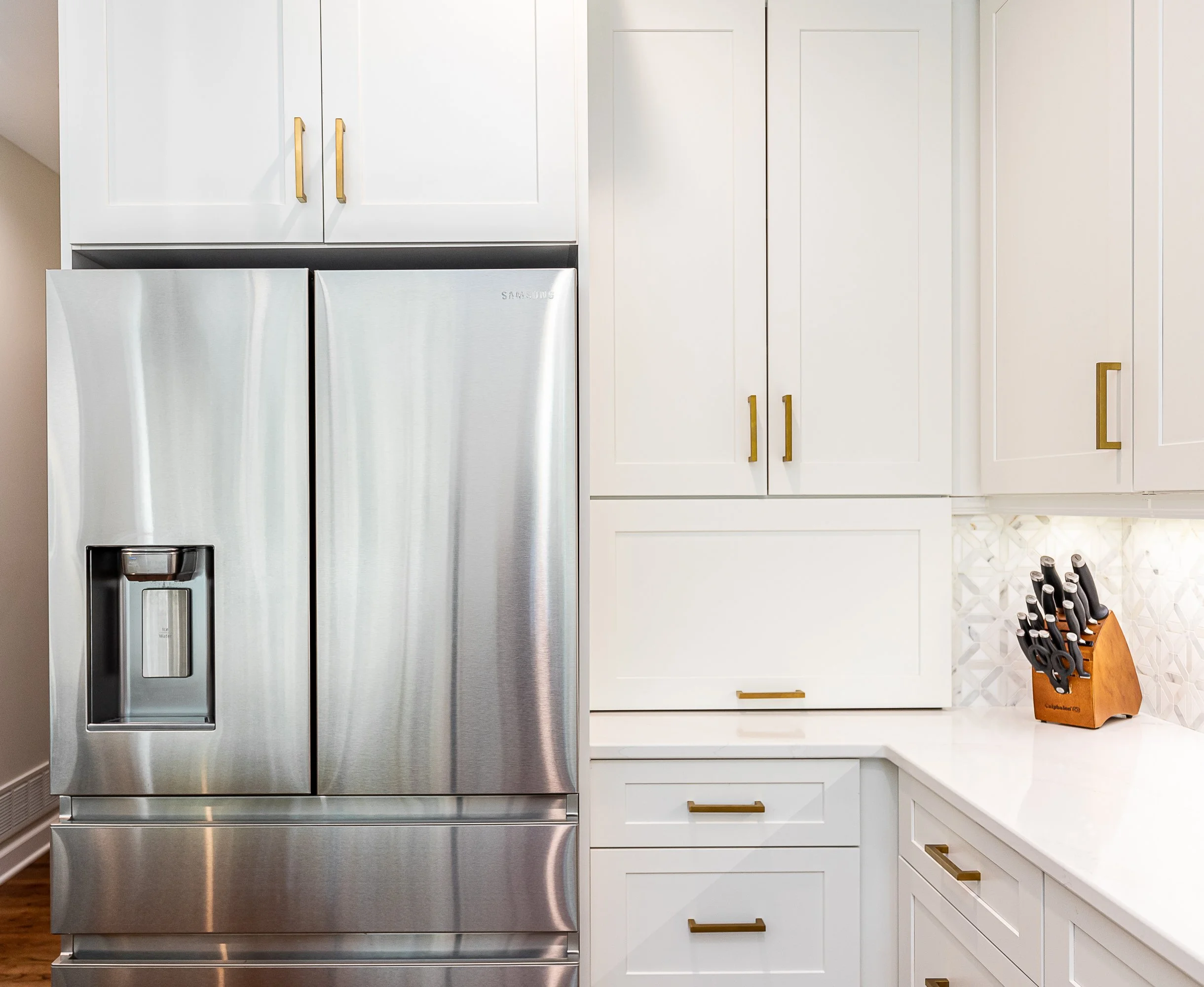 Stainless steel refrigerator in a modern kitchen with white cabinets and gold handles, and a knife block on the countertop.