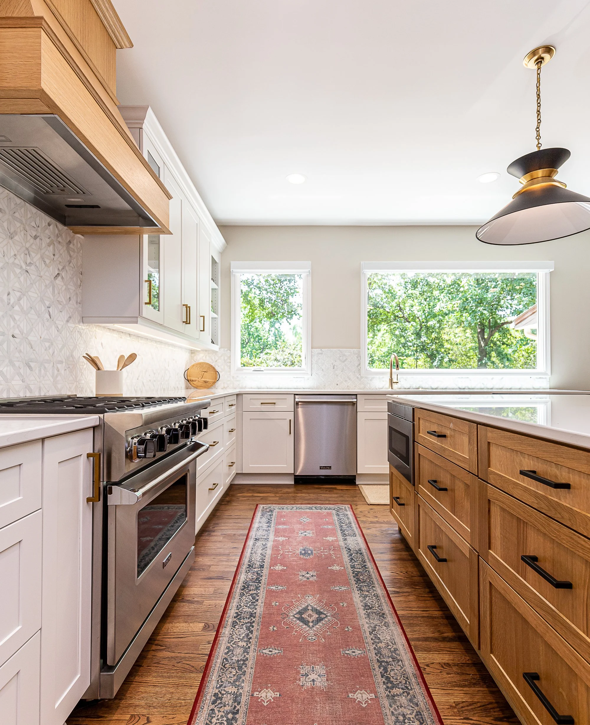 Modern kitchen with white and wooden cabinetry, stainless steel appliances, hardwood floor, and two large windows showing green trees outside.