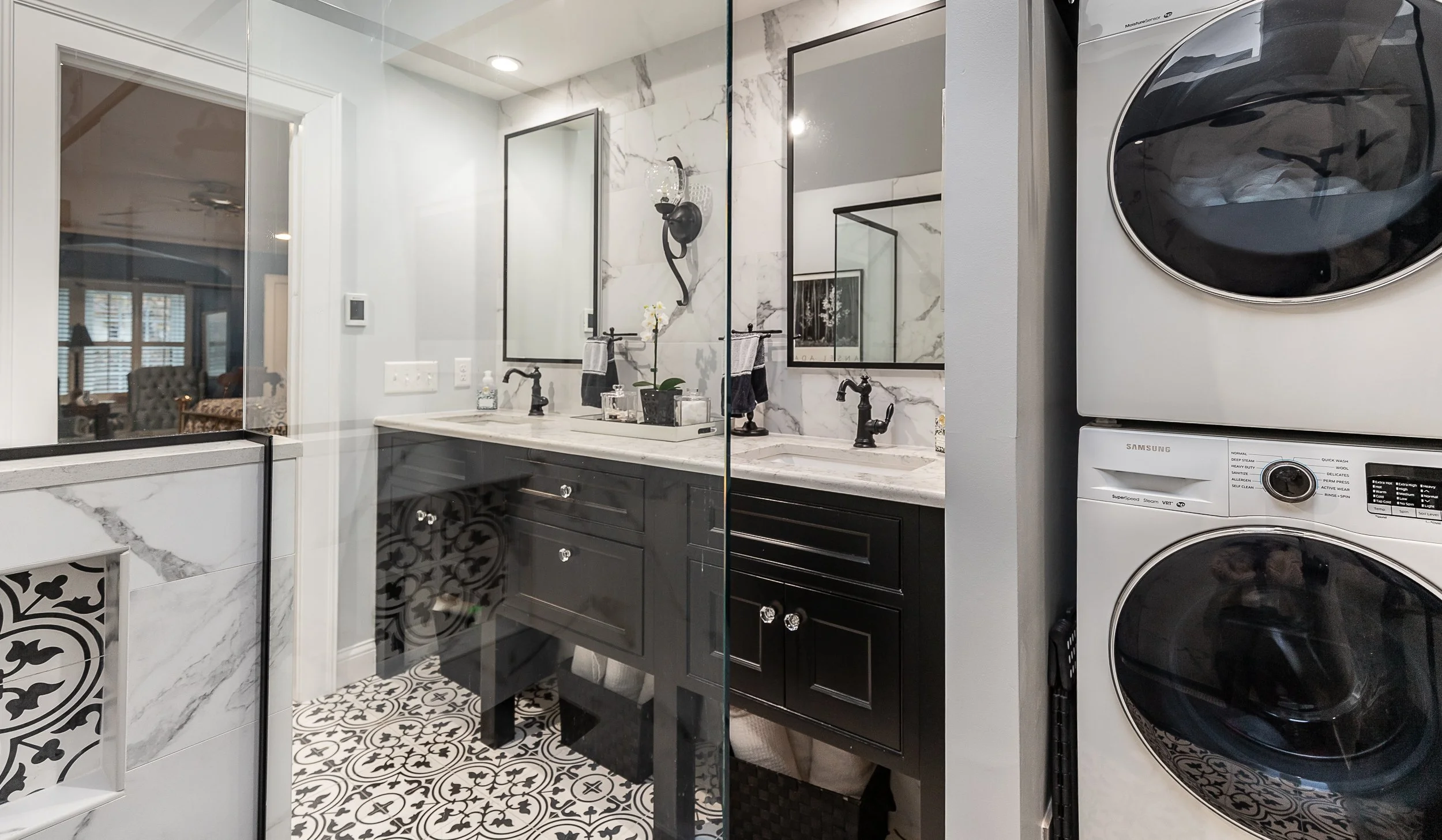 Bathroom with black vanity, double sinks, and mirrors, with decorative black and white patterned floor tiles, a glass shower door, and a stacked washer and dryer to the right.