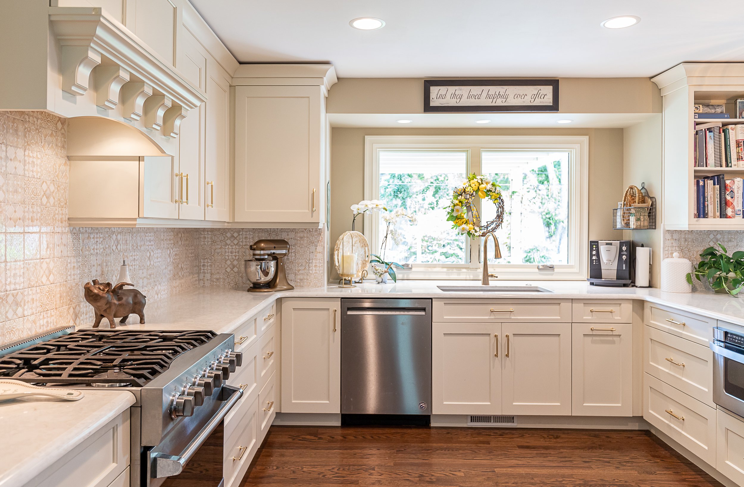 Beige kitchen with gold hardware, stainless steel appliances, a window above the sink with a wreath, and decorative items like a pig figurine and plants.