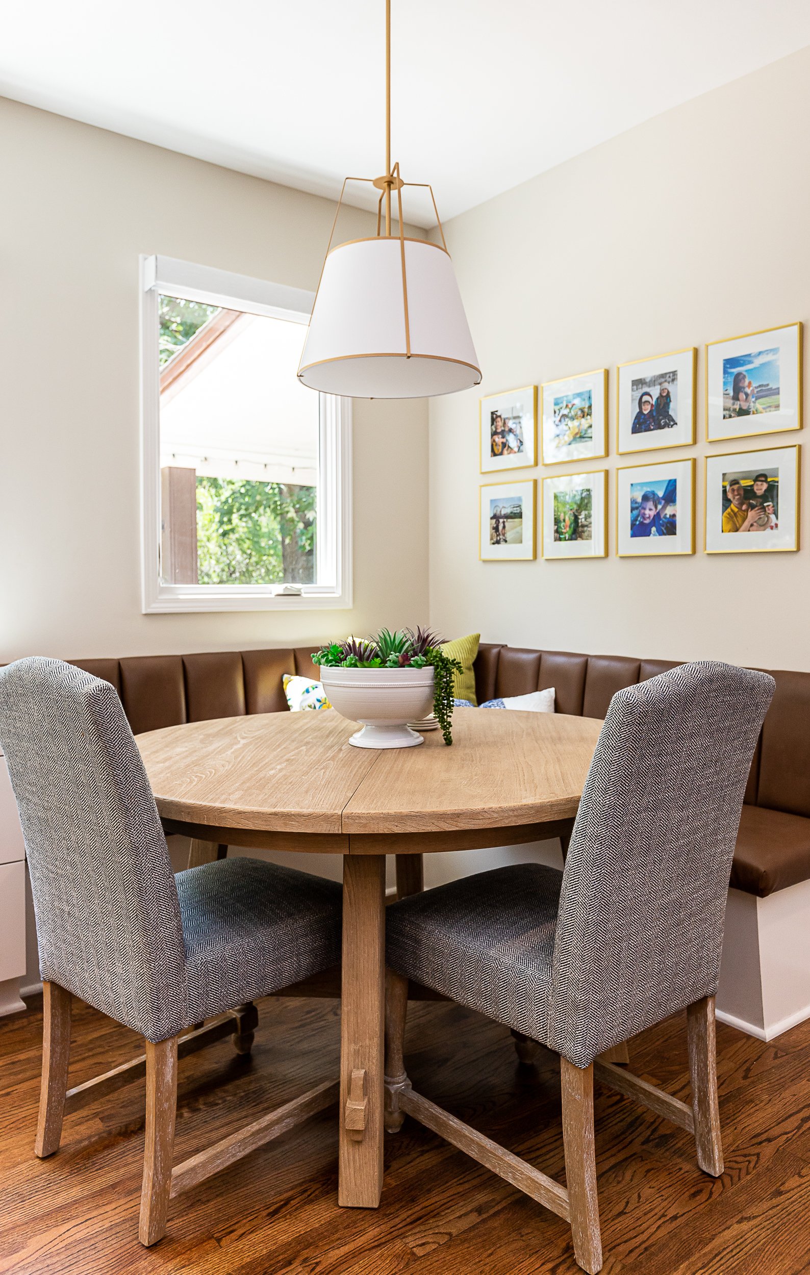 A round wooden dining table with four gray upholstered chairs around it. A white bowl with green plants is at the center of the table. Behind the table is a brown banquette with pillows. A window with a view of trees is on the left, and framed photographs are on the right wall. A large white pendant light hangs above the table.
