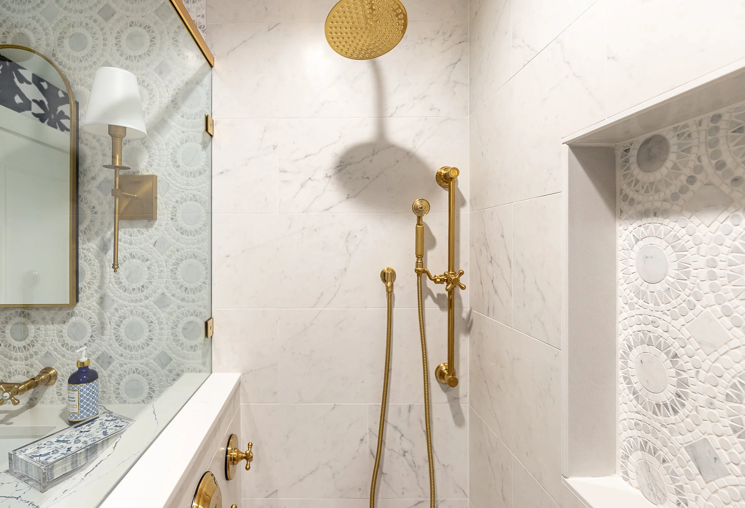 A bathroom shower with a gold shower head, handheld shower, and grab bar set against white marble-like tiles. There is a window with decorative tile framing a built-in shelf, a mirror, and a wall-mounted lamp with a white shade.
