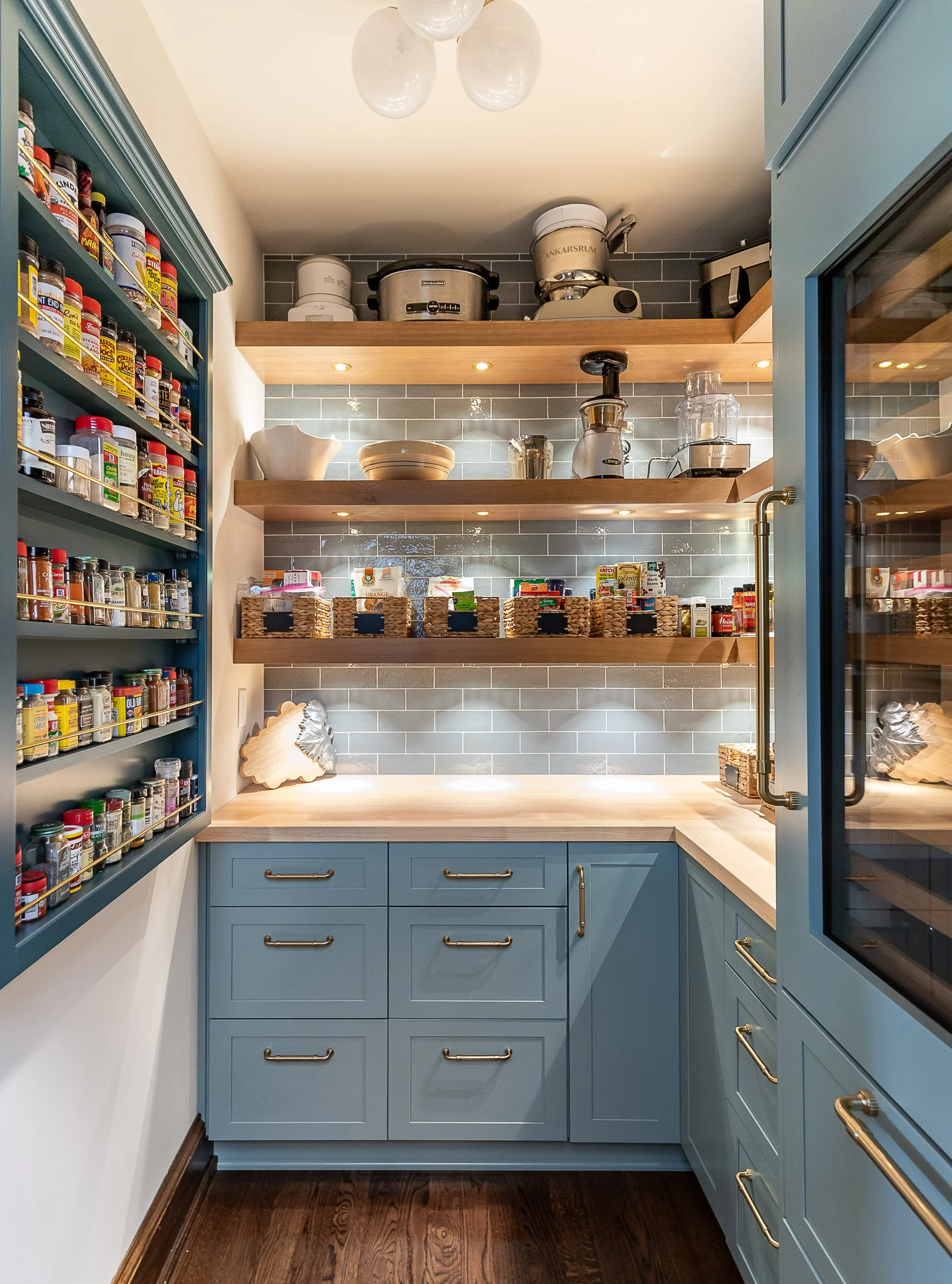 A cozy kitchen with blue cabinets, wooden open shelves, a white countertop, and a dark wood floor. The shelves hold various kitchen appliances, bowls, spices, and food items. The kitchen has a modern design with gray tiled backsplash and warm lightin