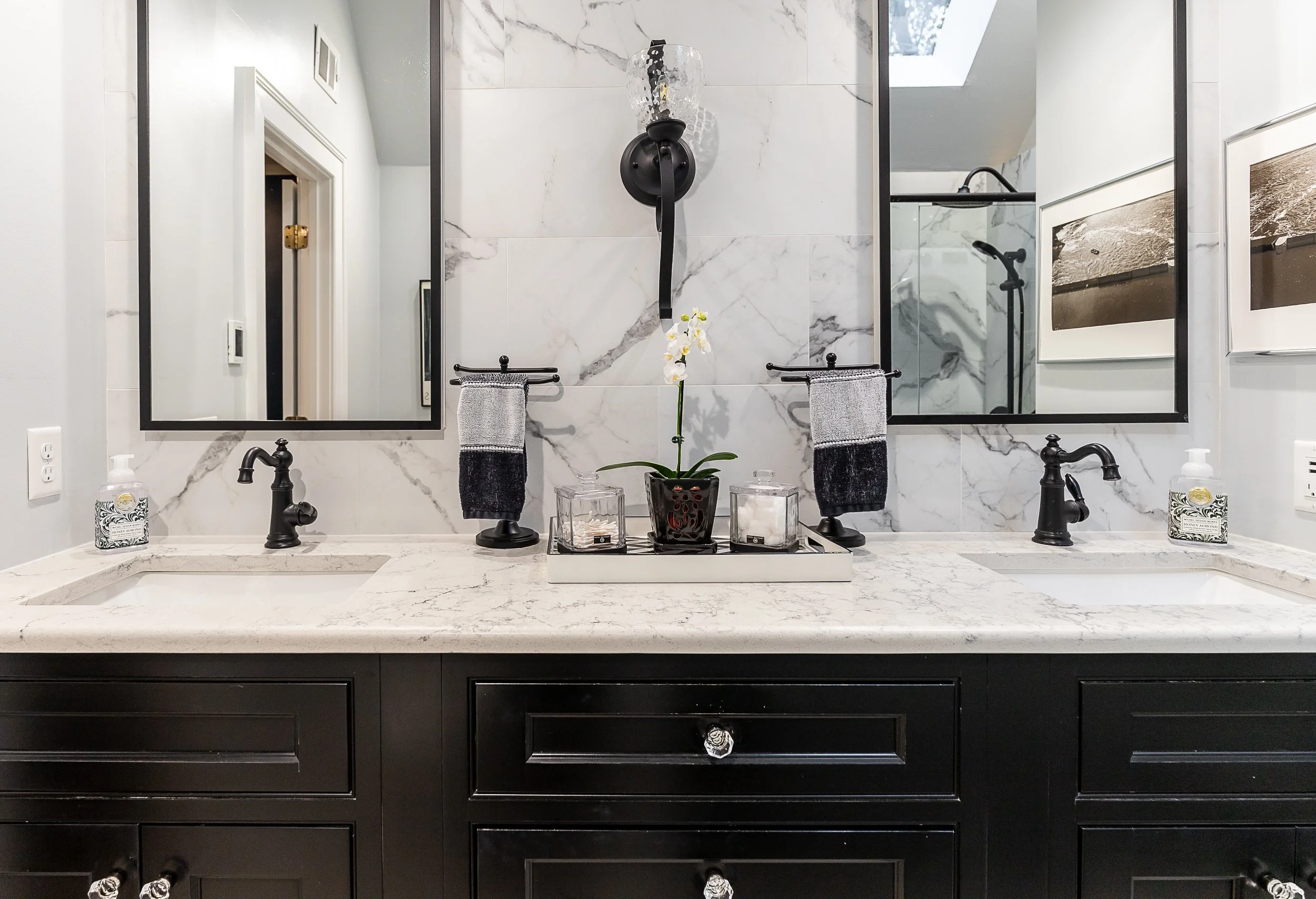 A modern bathroom vanity with a double sink, black fixtures, and a quartz countertop, featuring a large mirror, a potted orchid, decorative candles, and two hand towels.