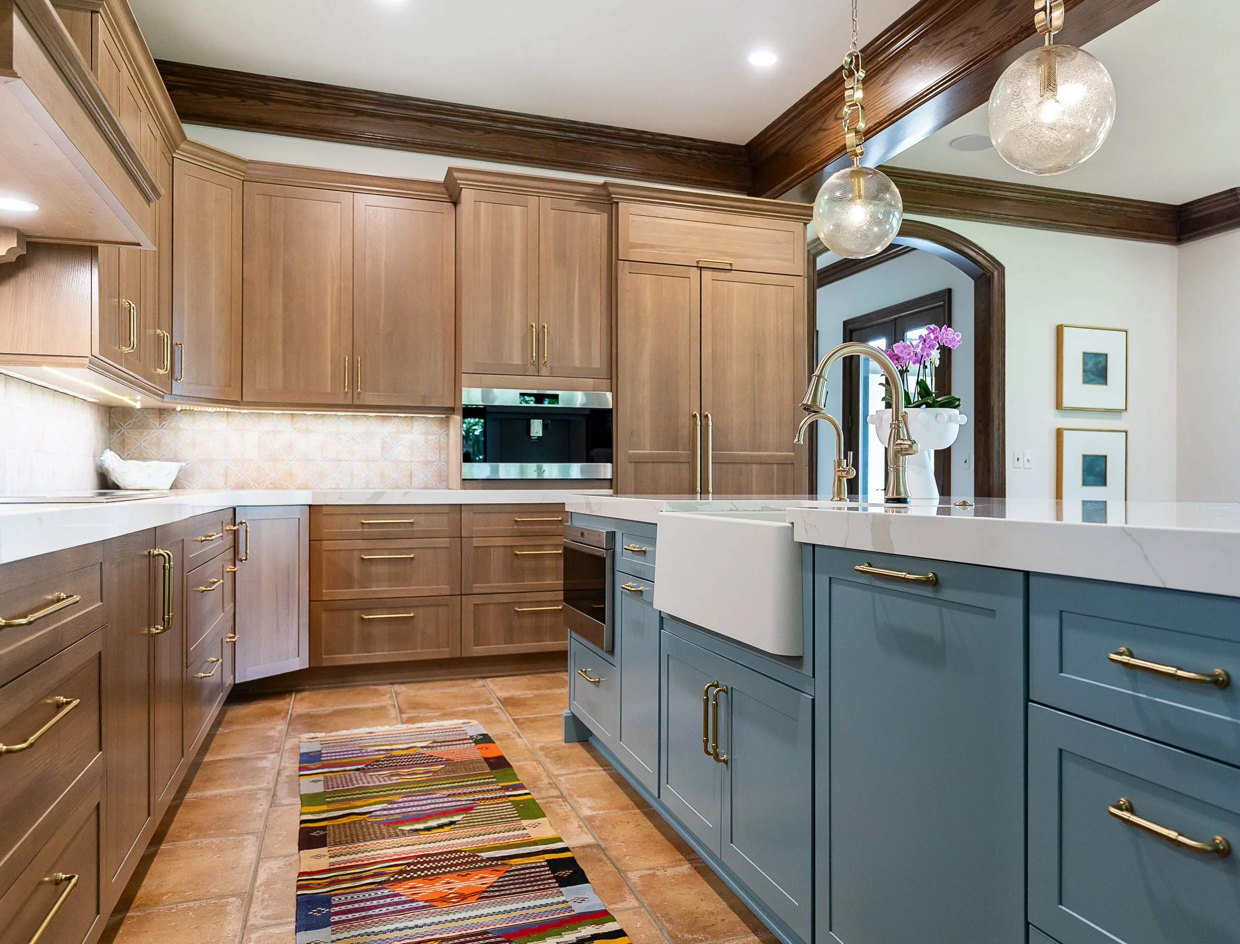 A spacious kitchen with light wood cabinets, a white marble island, gold handles, hanging globe pendant lights, a orchid plant, and a colorful striped rug on a tiled floor.