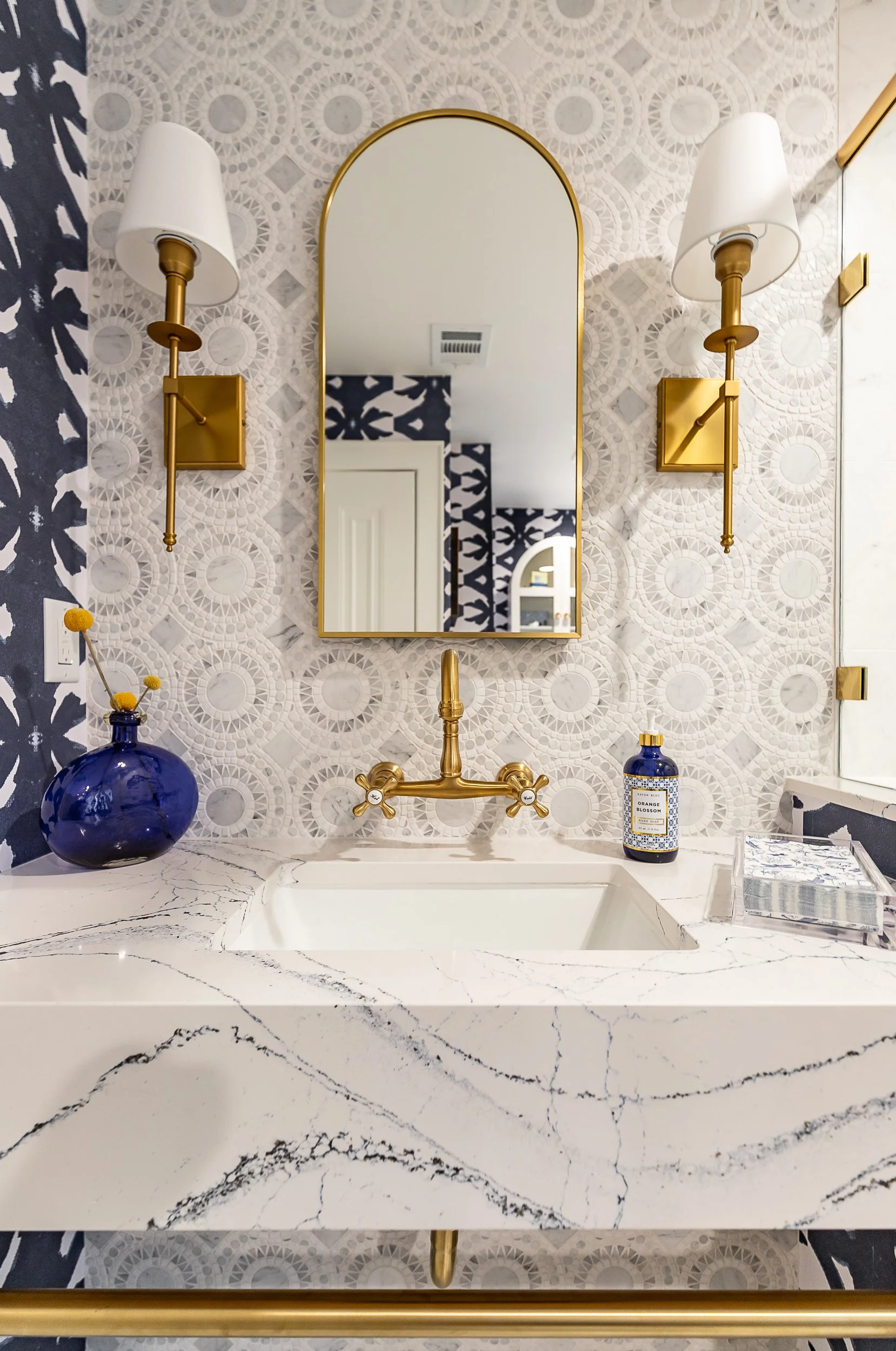 Bathroom with marble-looking countertop, gold fixtures, a mirror, blue vase with yellow flowers, and decorative wallpaper.
