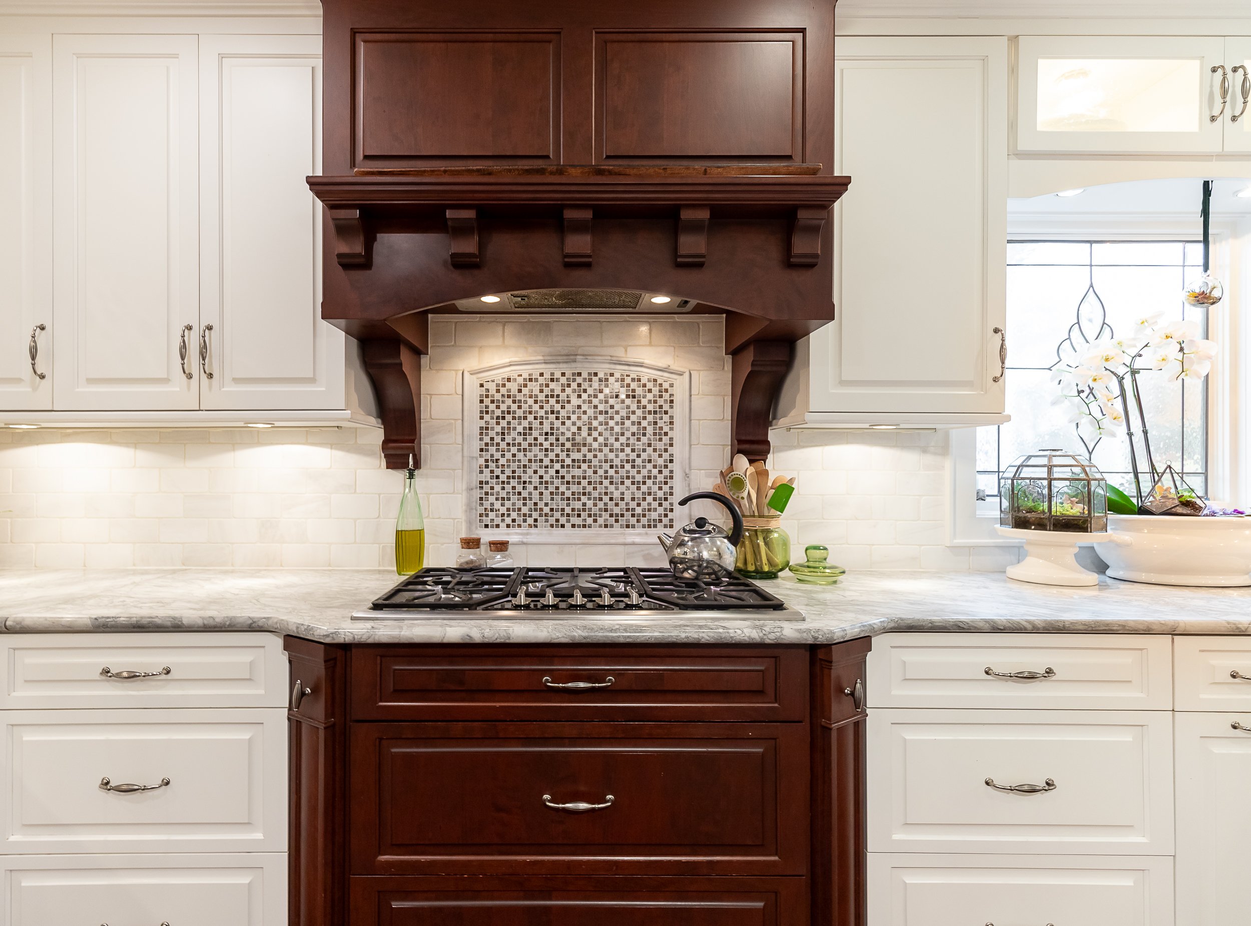 Kitchen with white cabinets, a marble-like countertop, and a wooden range hood over a gas stove. There are small plants in a glass terrarium and a white flowerpot on the countertop near a window.