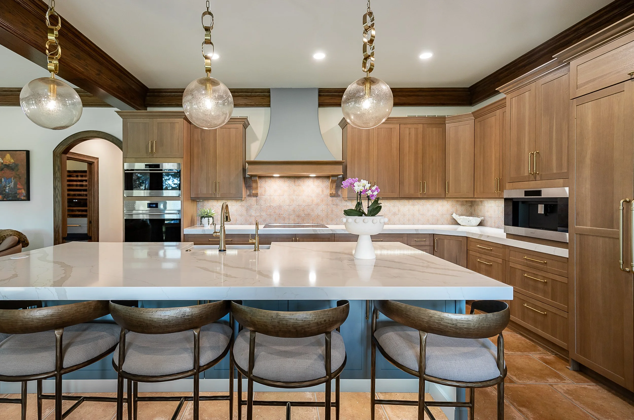 A modern kitchen with wooden cabinets, a large white island, hanging globe pendant lights, and a vase with purple and white orchids on the countertop.