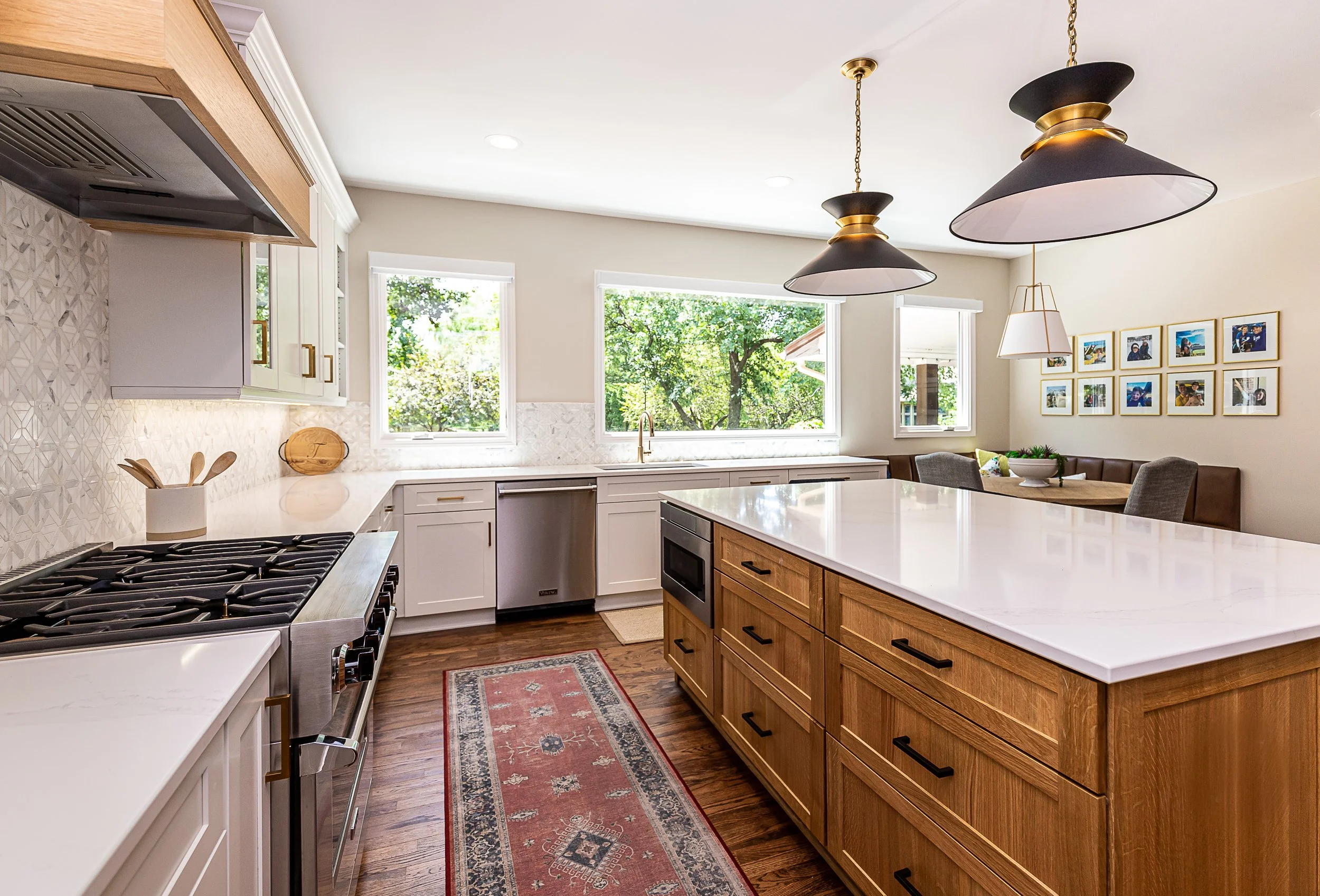 Modern kitchen with white cabinets, a large island with a marble countertop, wooden lower cabinets, a gas stove, and large windows showing greenery outside.