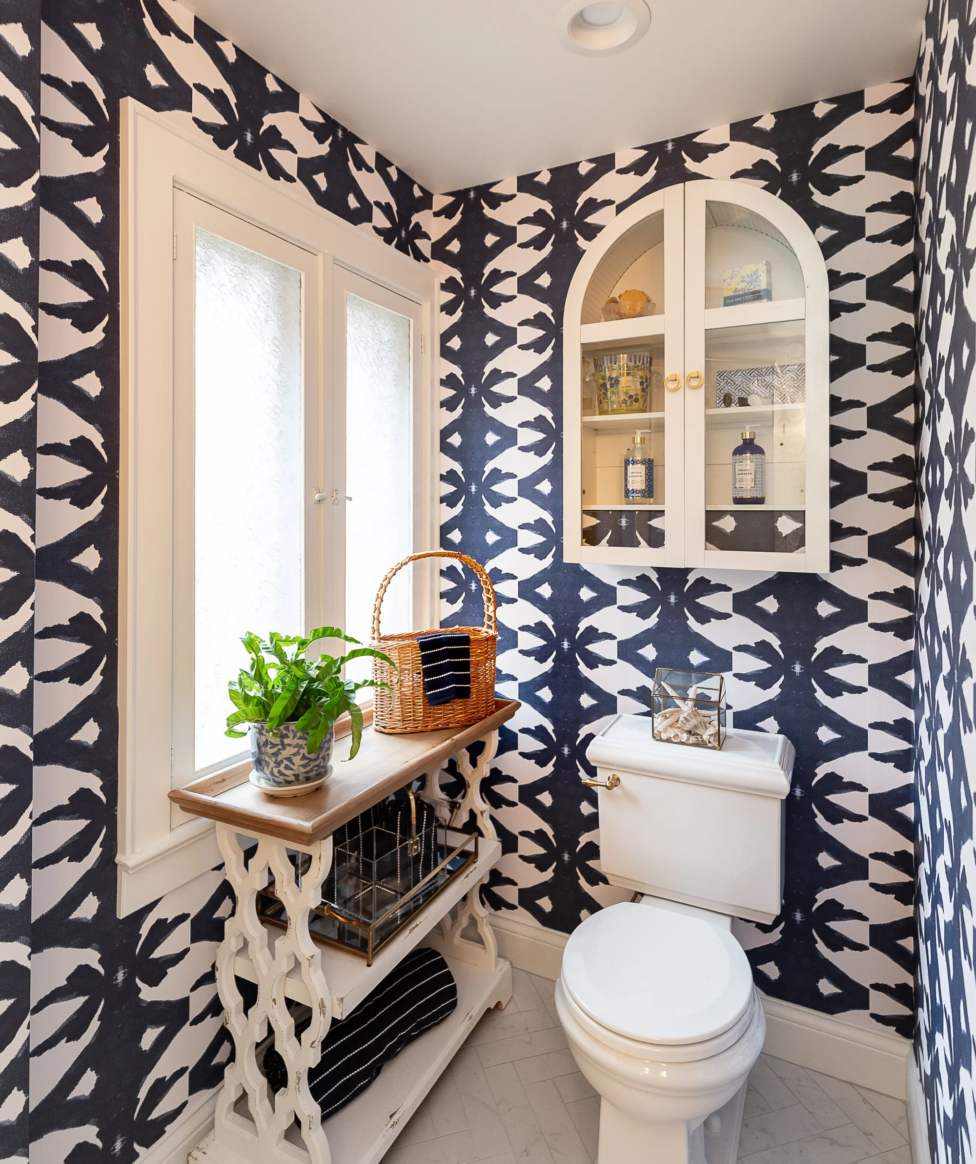 Small bathroom with a geometric patterned wallpaper, a white toilet, a window, a white wall-mounted cabinet with arched top and glass doors, a white ornate table with a plant in a ceramic pot and a wicker basket with a clothing item, and decorative items on shelves.
