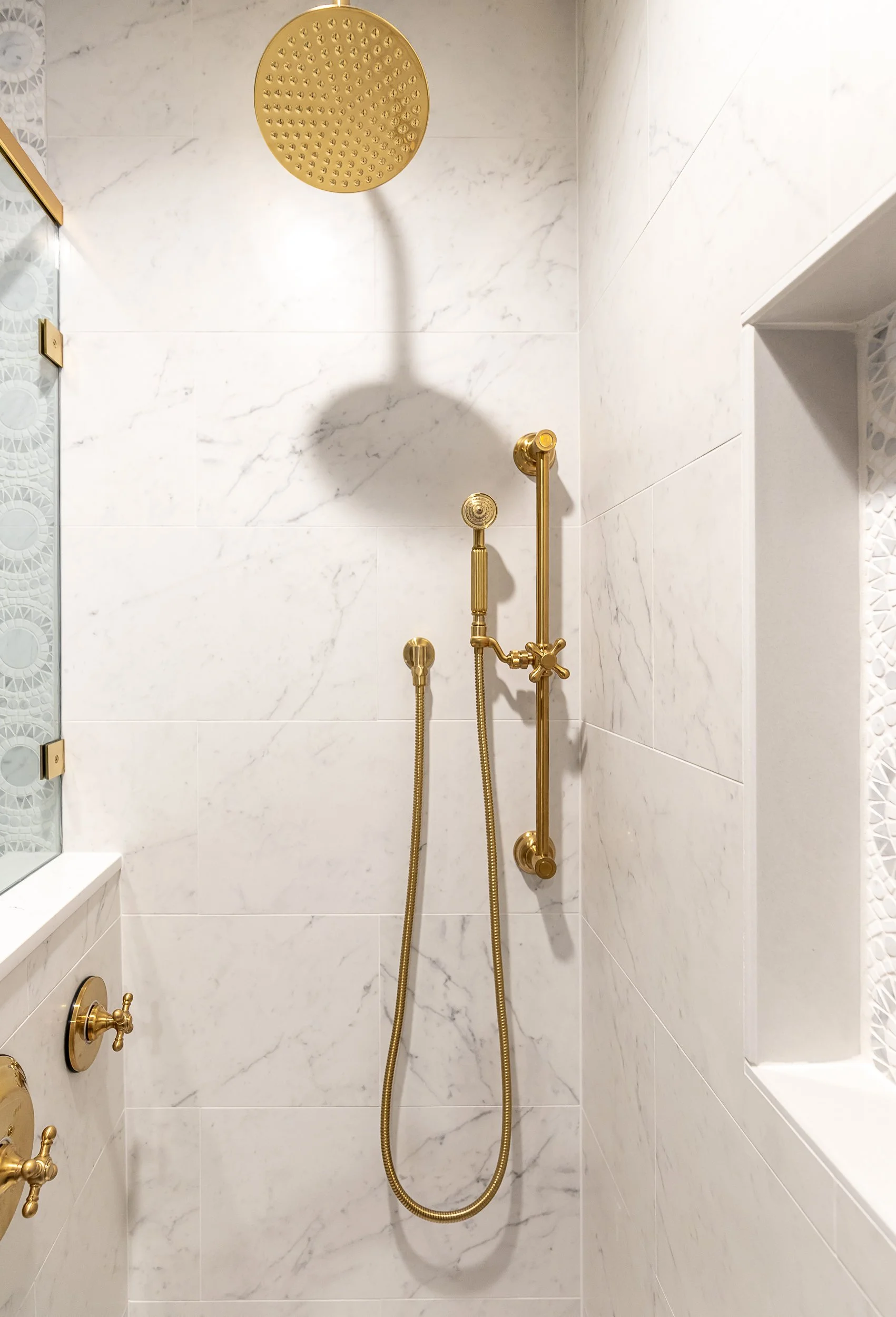 A modern shower with white marble-looking tiles, gold fixtures, a large round rain showerhead, and a hand-held shower head with a flexible hose.