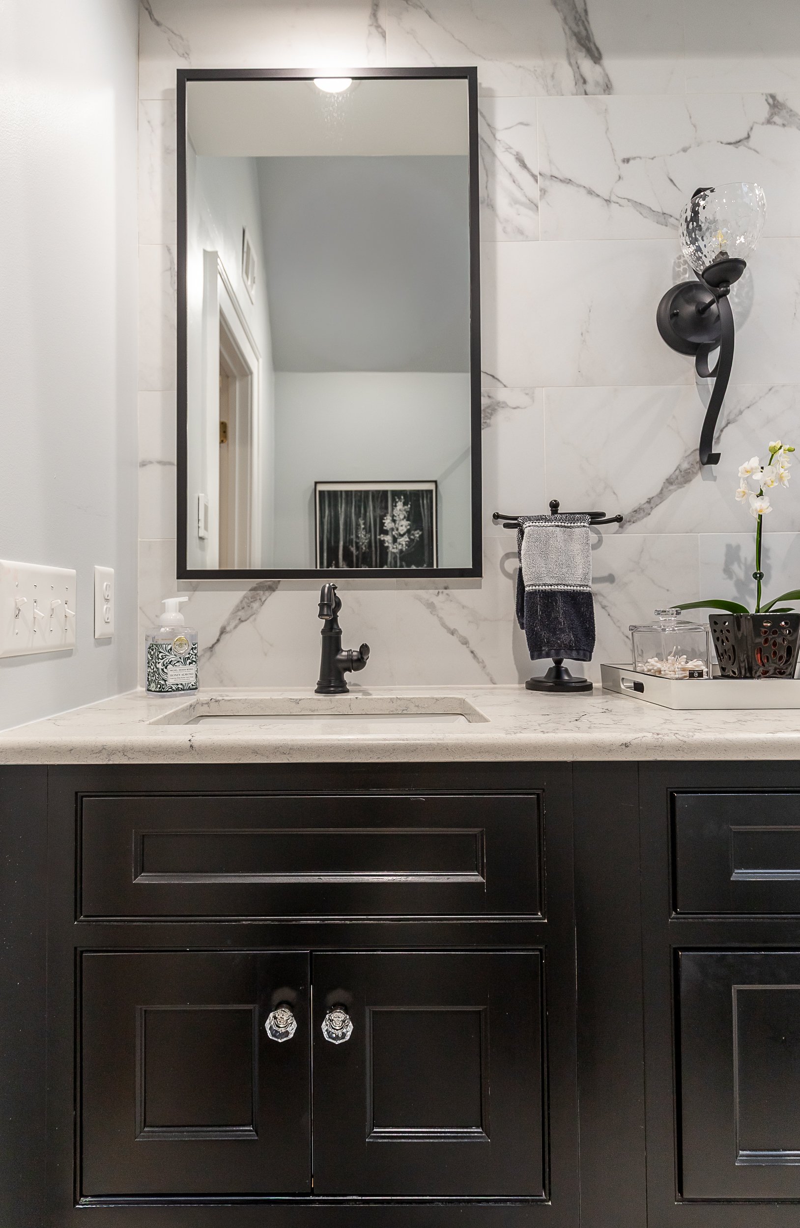 Bathroom vanity with a large mirror, black cabinet, white marble countertop, black faucet, hand sanitizer, towel holder with two gray towels, wall-mounted light fixture, and decorative items including a potted orchid plant and a tray.