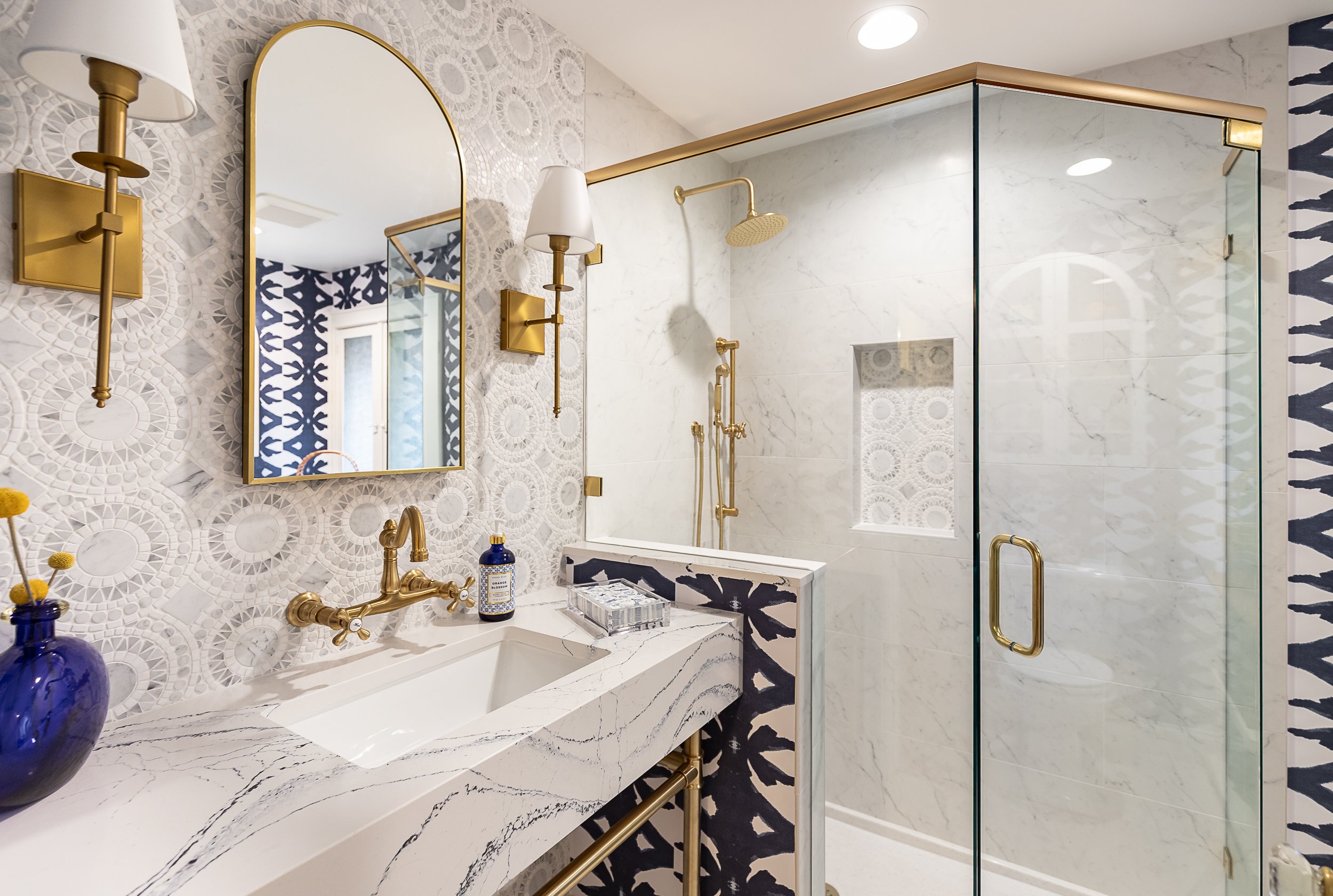 Modern bathroom with white marble sink and shower, gold fixtures, patterned wallpaper, and a mirror.