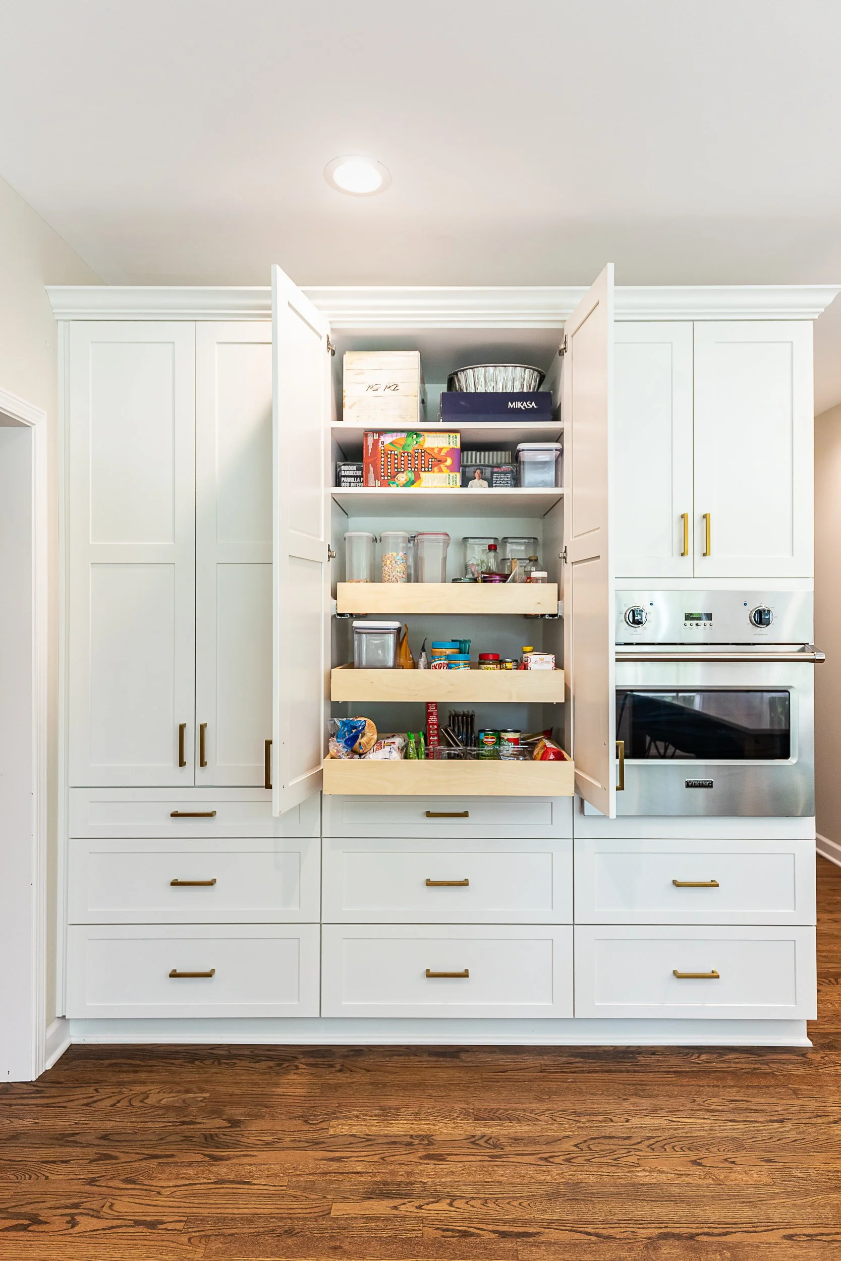 Open pantry in a kitchen with white cabinets, wooden shelves, and a built-in oven. The pantry contains canned goods, jars, and boxes of food. The kitchen has a hardwood floor.