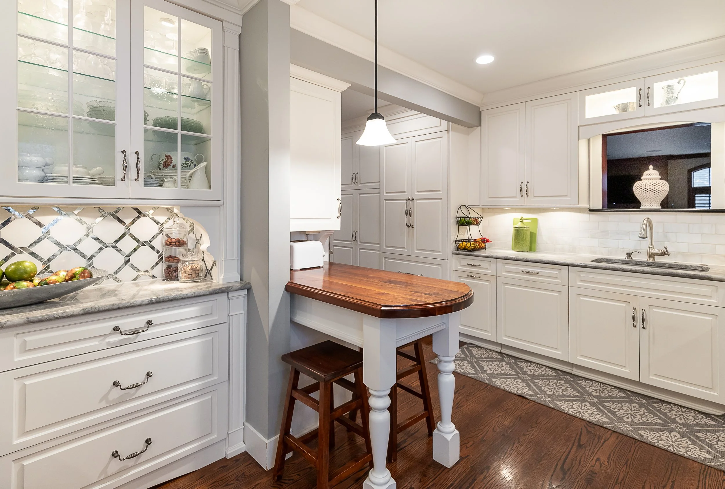 A cozy kitchen with white cabinets, a countertop, and a wooden breakfast bar. Glass-front cabinets display dishes, and a patterned backsplash is visible. The floor is wooden and a pendant light hangs over the breakfast area.